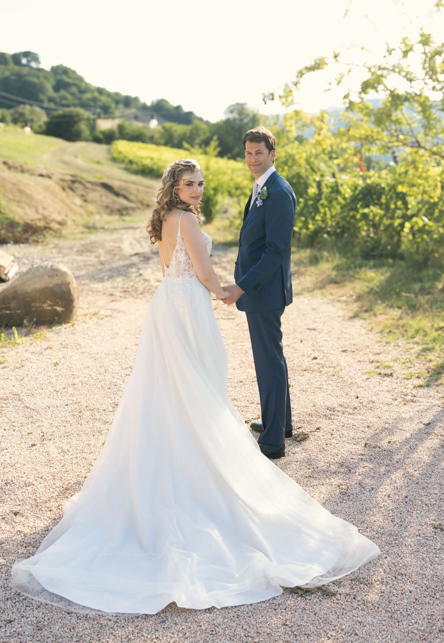 A bride in a flowing white gown walking along a dirt path in a vineyard setting.