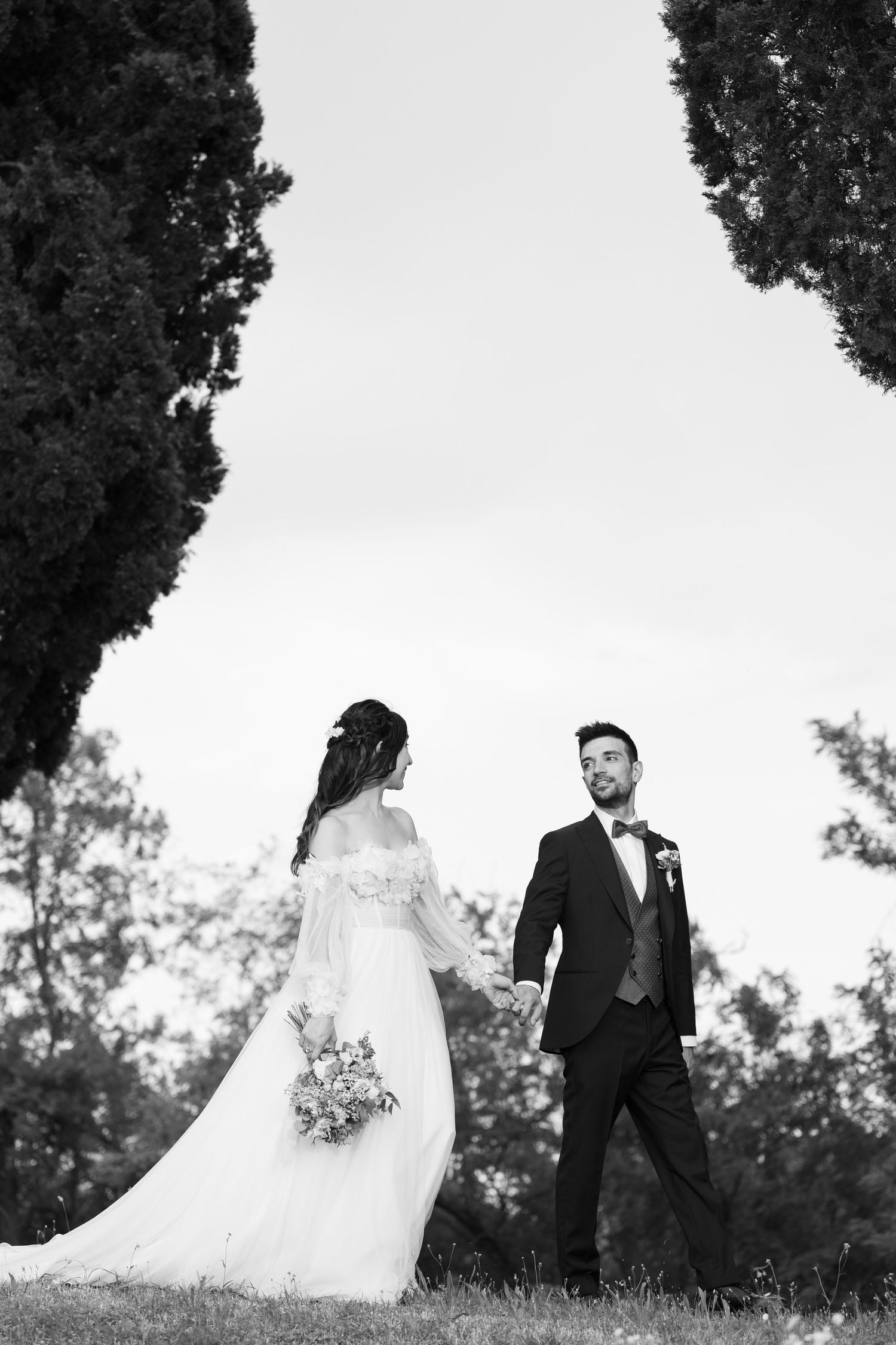 Black and white photo of newlyweds standing between cypress trees in an outdoor setting.