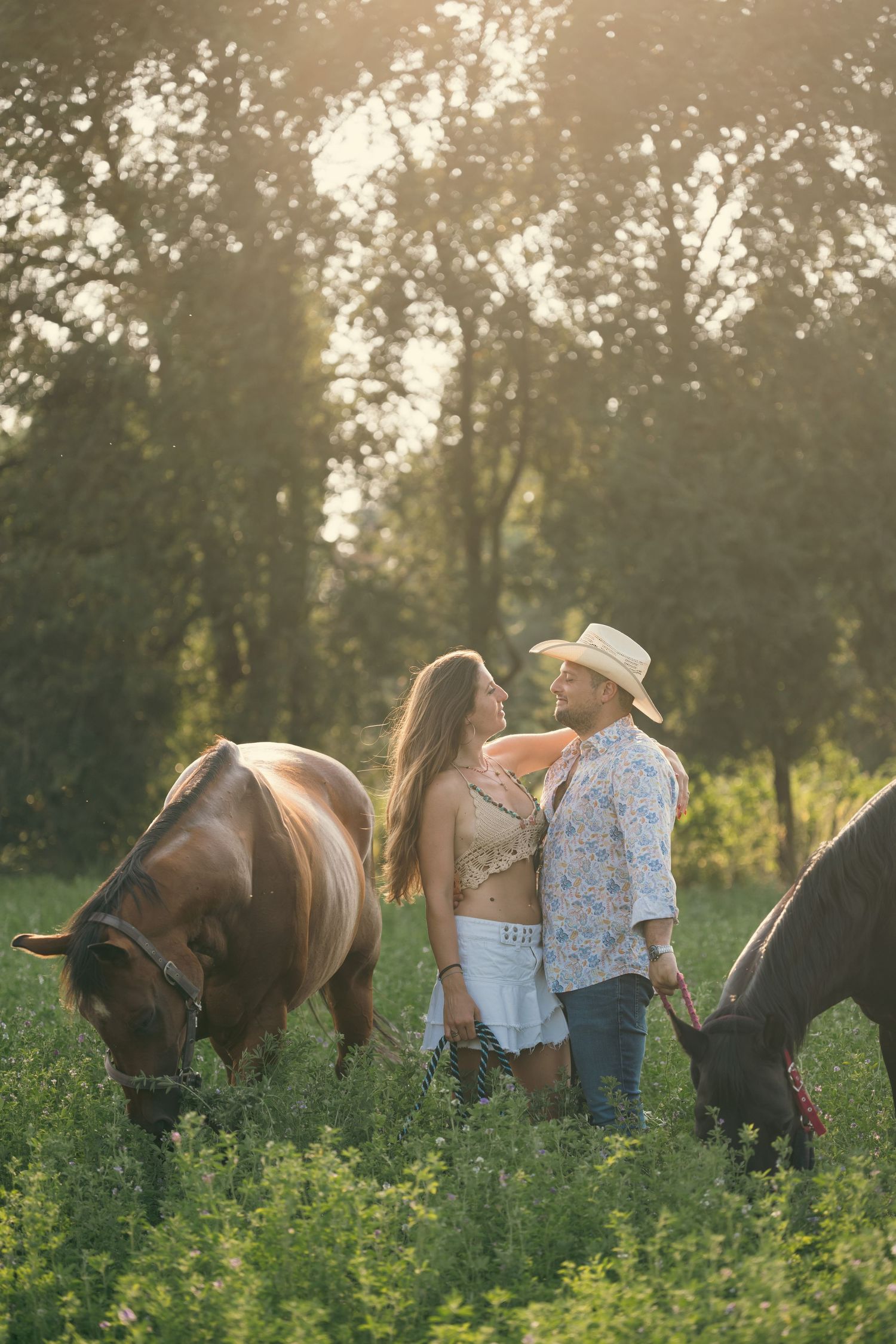 Couple posing for romantic sunset photos in field with horses grazing nearby.