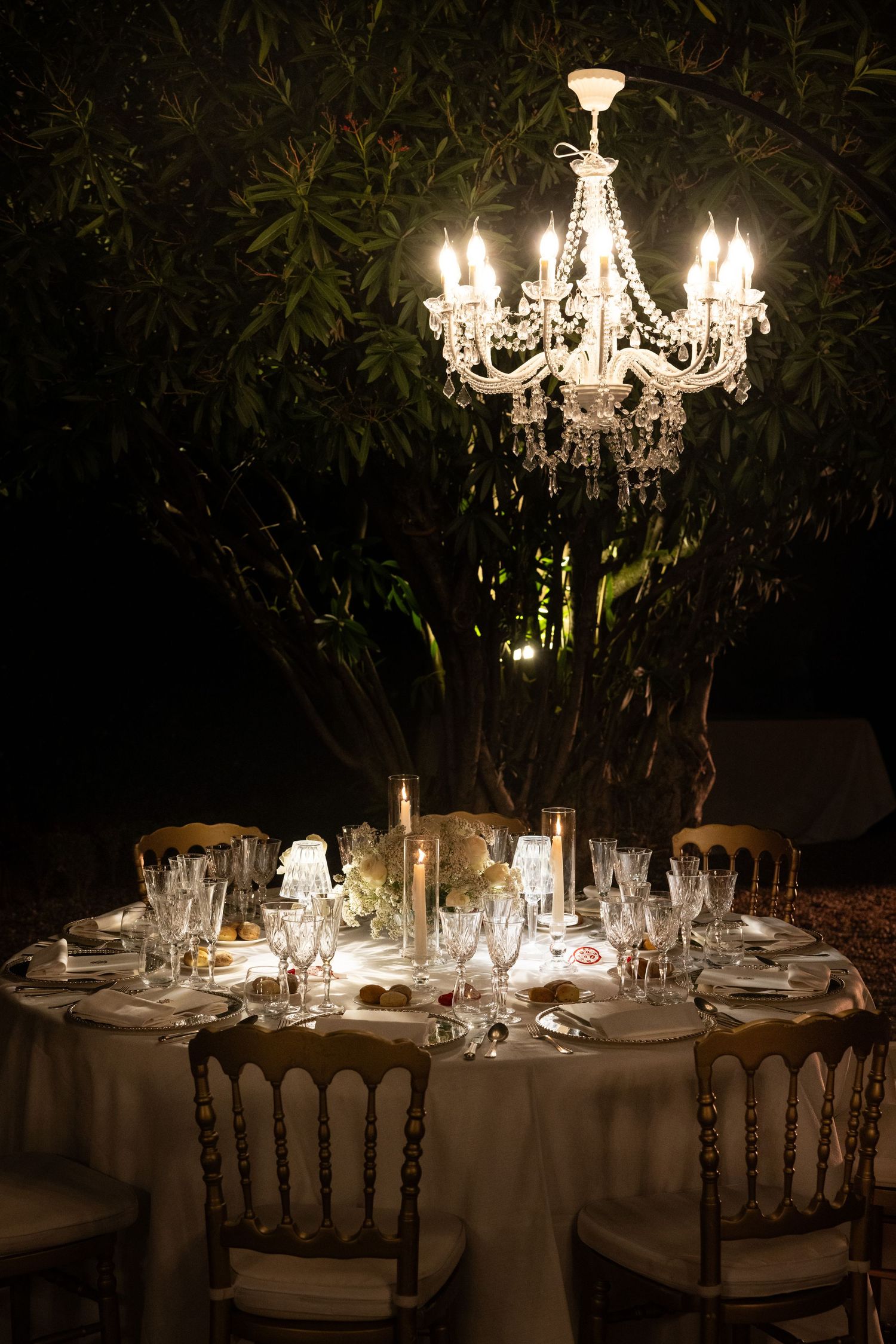 Elegant outdoor dining tables under crystal chandeliers with garden backdrop at night.