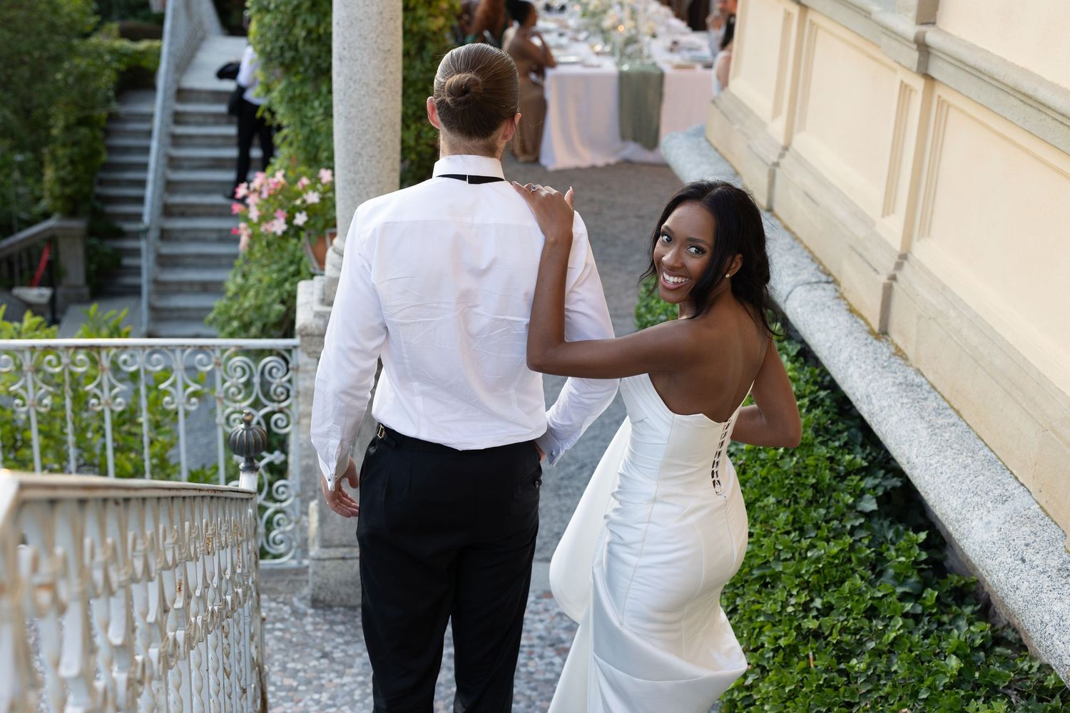 A couple in white outfits dances together on a garden terrace during what appears to be a wedding celebration.