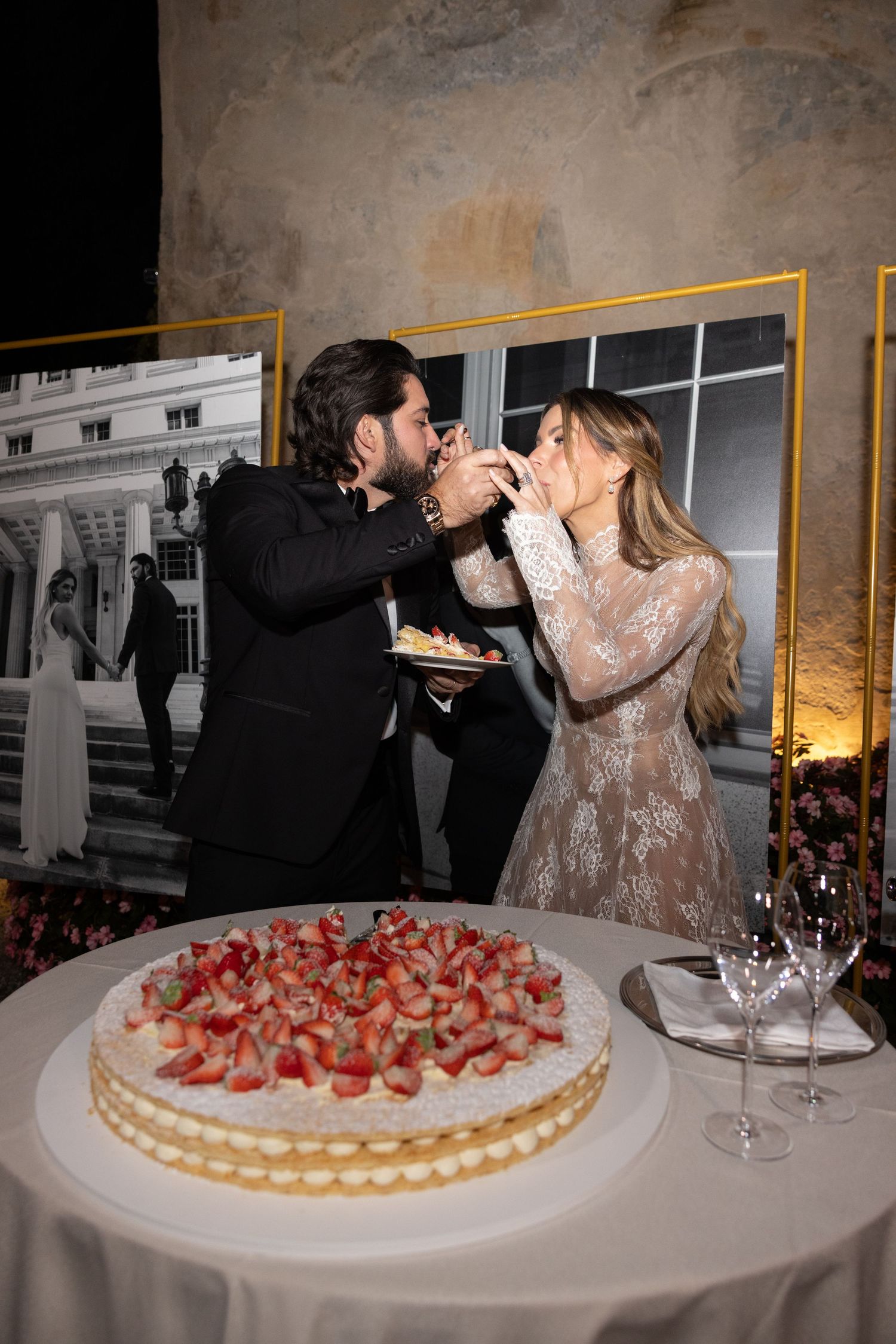 Couple shares champagne toast while cutting a strawberry-topped wedding cake at their reception celebration.