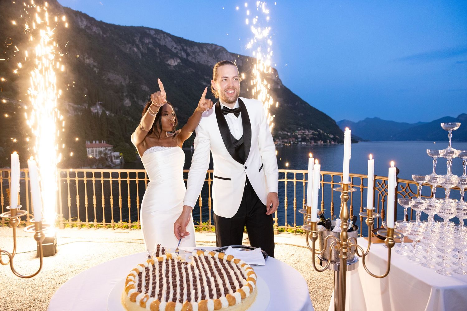 Elegant wedding reception setup at dusk with sparklers and candlelit tables along a waterfront mountain venue.