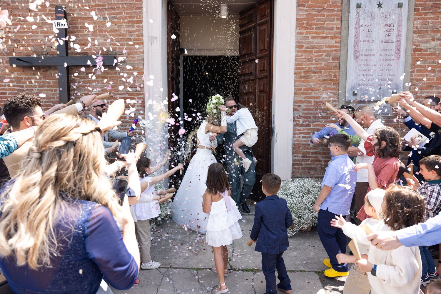 A joyful crowd throws white confetti during an outdoor wedding celebration.