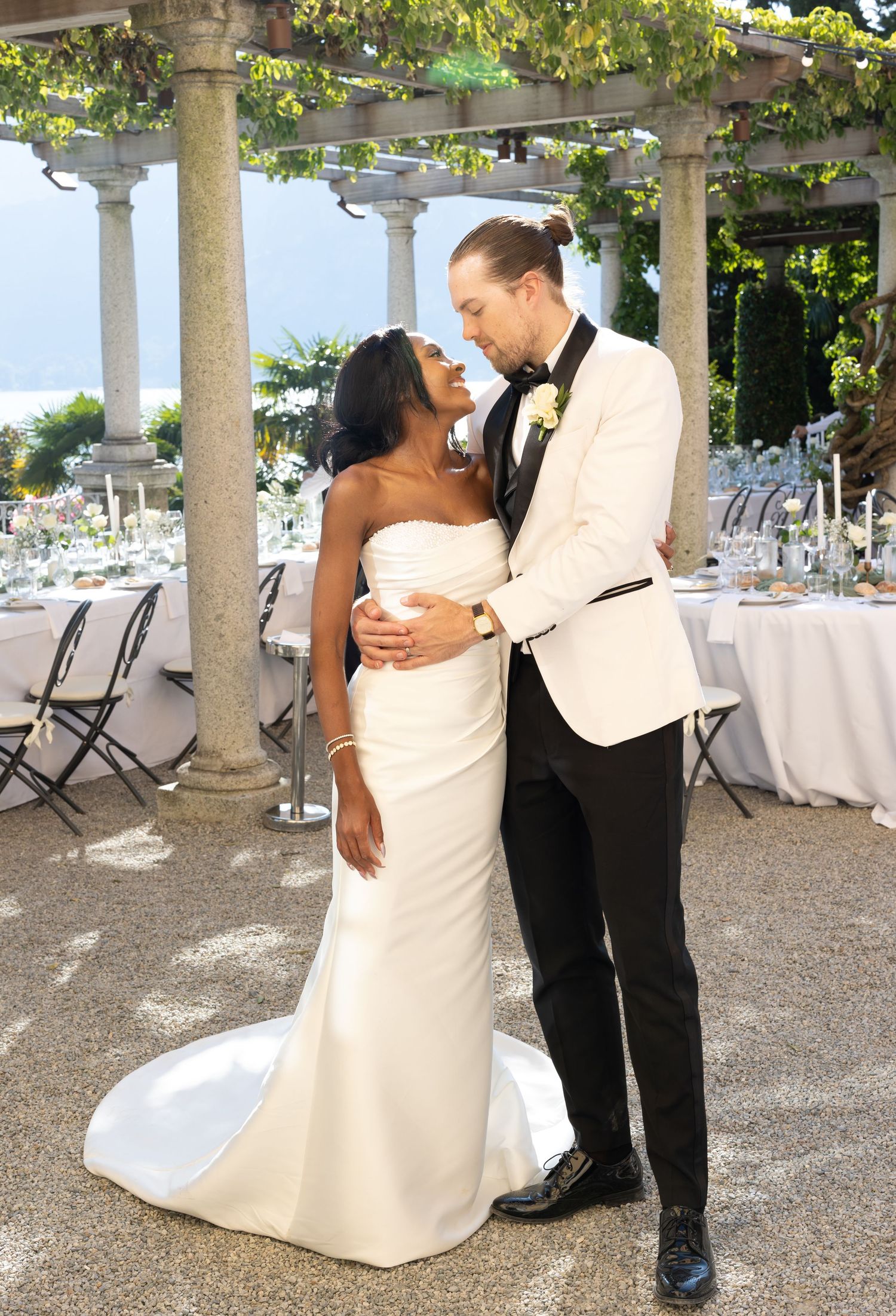 A romantic wedding couple embraces under a pergola decorated with flowers at an outdoor reception venue by the sea.