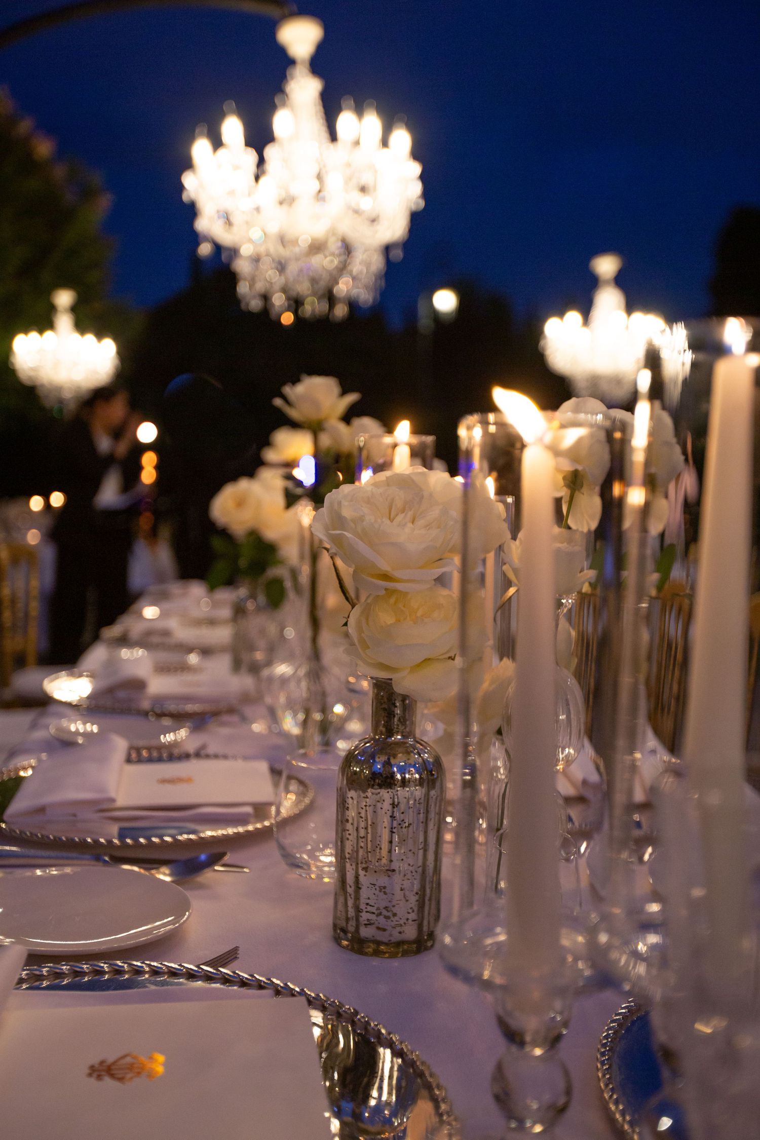 Romantic table setting with white roses and crystal chandeliers for evening wedding reception.