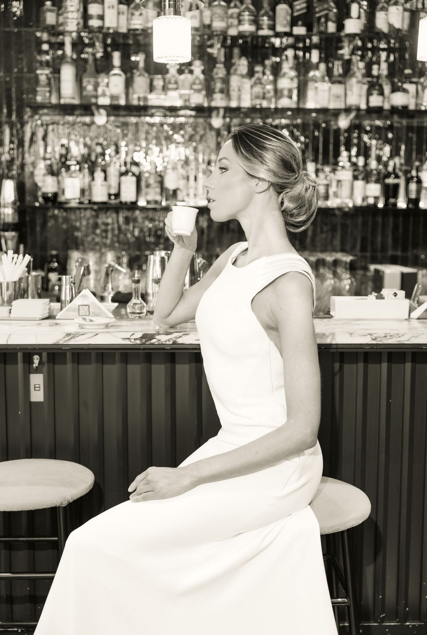 Black and white photo series of a wedding dress at an upscale bar with bottle displays.