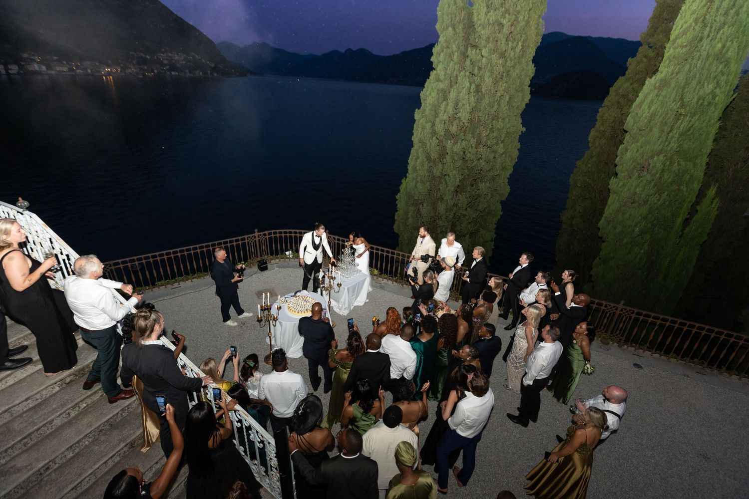 Wedding guests dance and celebrate during an outdoor evening reception with tall hedges in the background.