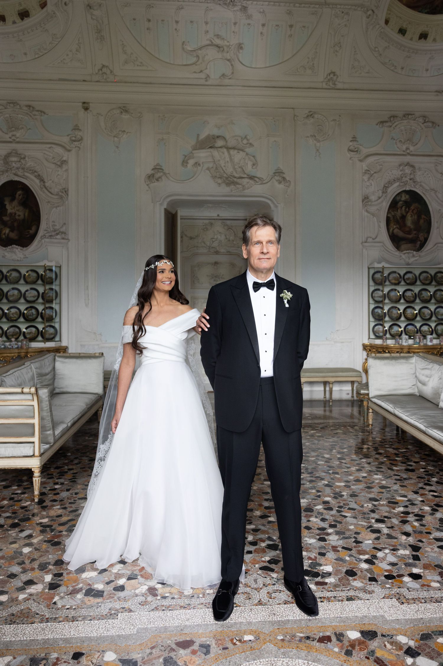 Newlyweds stand together in a grand room featuring ornate white walls and decorative ceiling paintings.