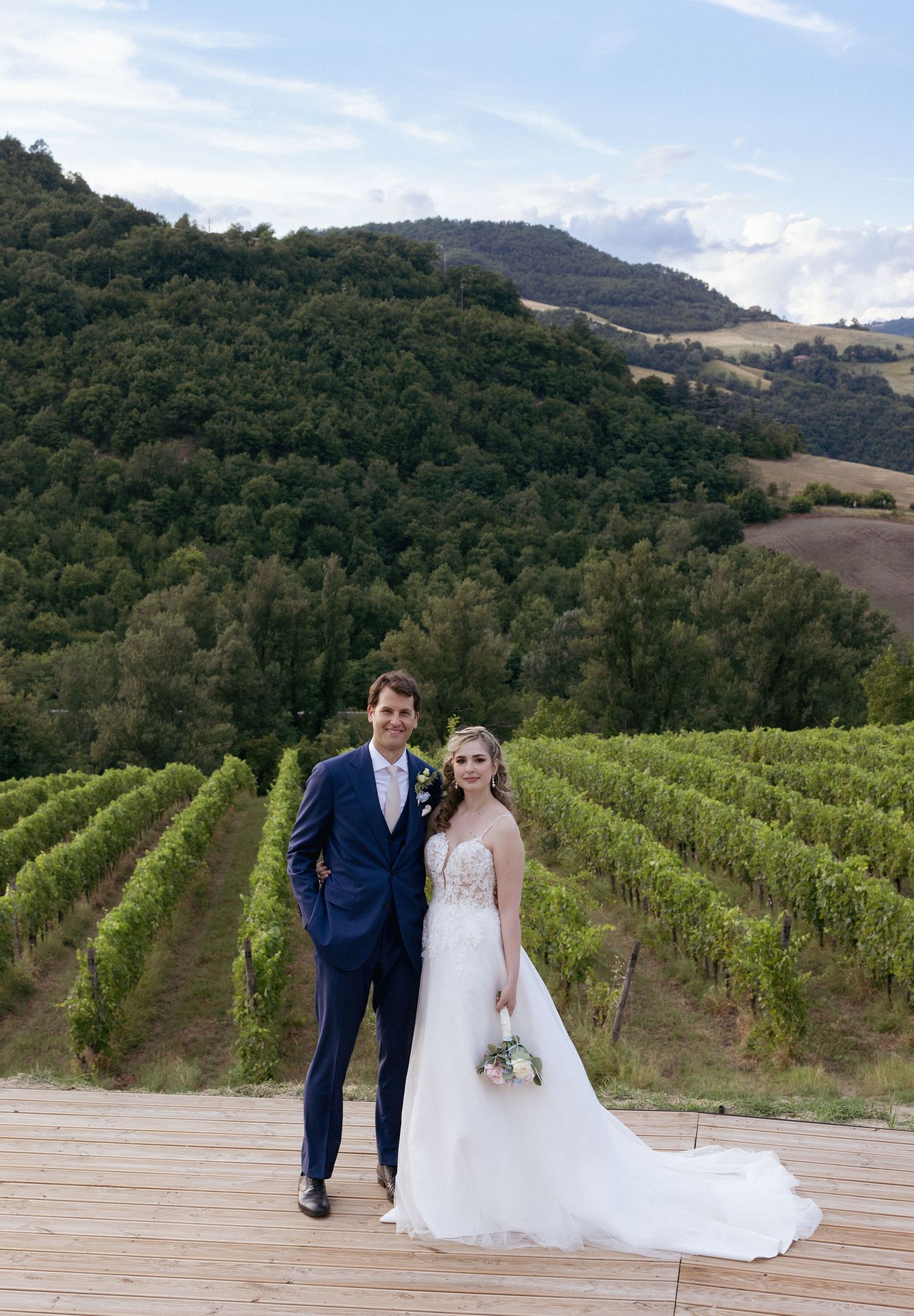 Newlyweds posing together on a wooden deck overlooking rolling vineyard hills.