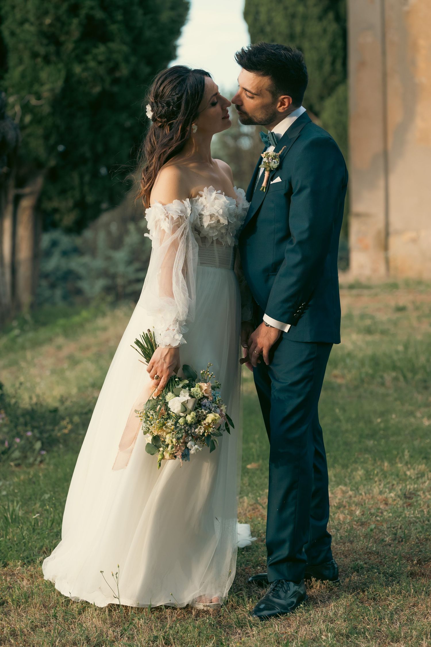 A romantic wedding couple embraces in a garden setting with the bride holding a delicate bouquet of flowers.