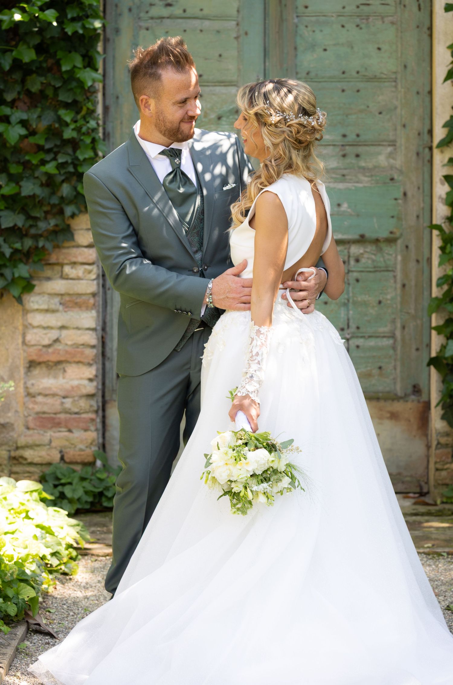 Newlyweds pose in front of vintage green doors with bride holding a delicate floral bouquet.
