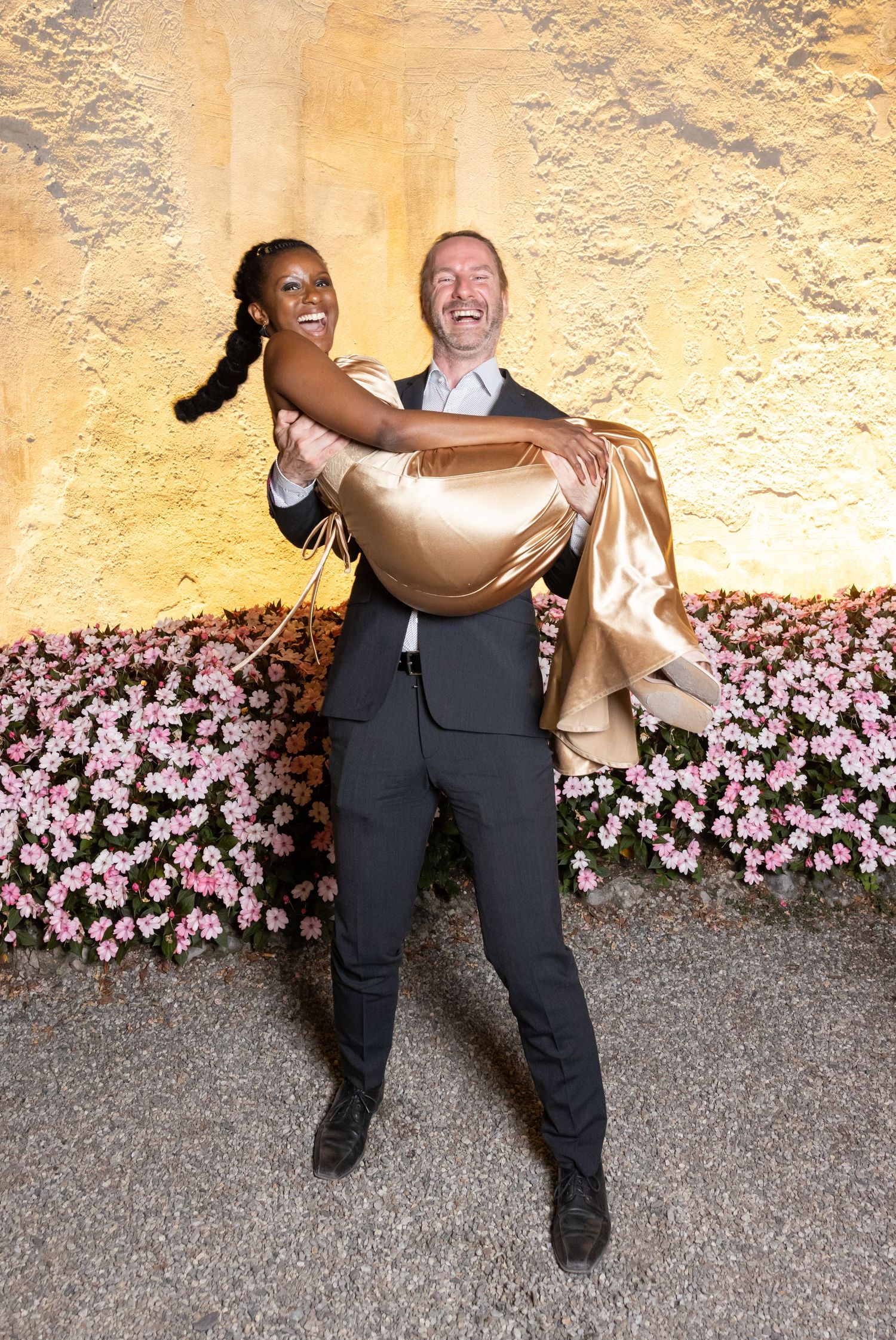 A couple shares a joyful moment against a yellow wall with pink flowers, as one person carries the other in their arms.