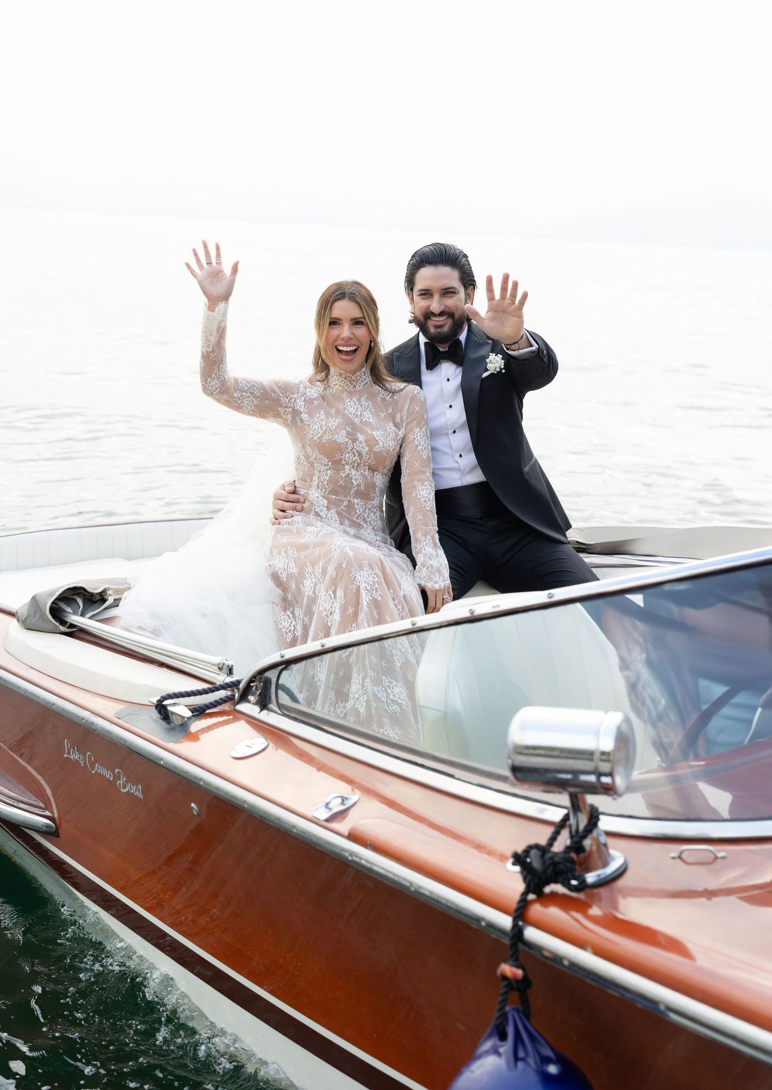 Newlyweds wave from a classic wooden boat on the water, with the bride in lace and groom in black tie.