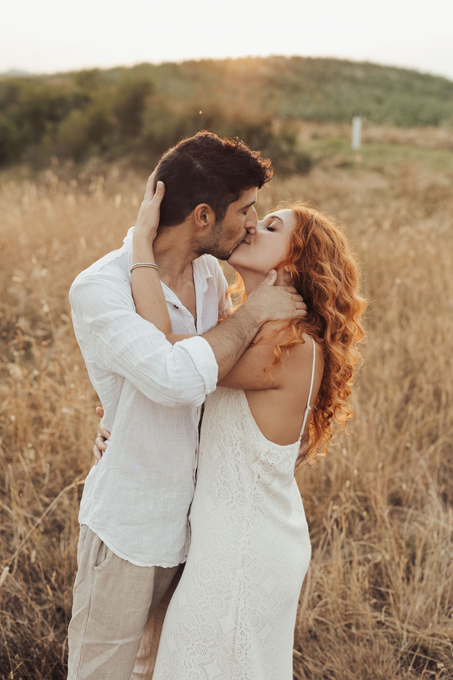 Romantic couple in white outfits sharing intimate moments in golden field at sunset.
