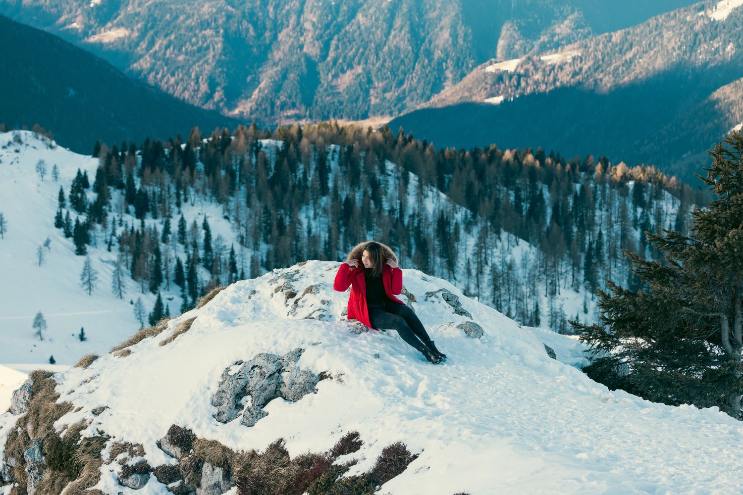 Skier in red jacket rests on snowy mountain peak with dramatic mountain vista in background.