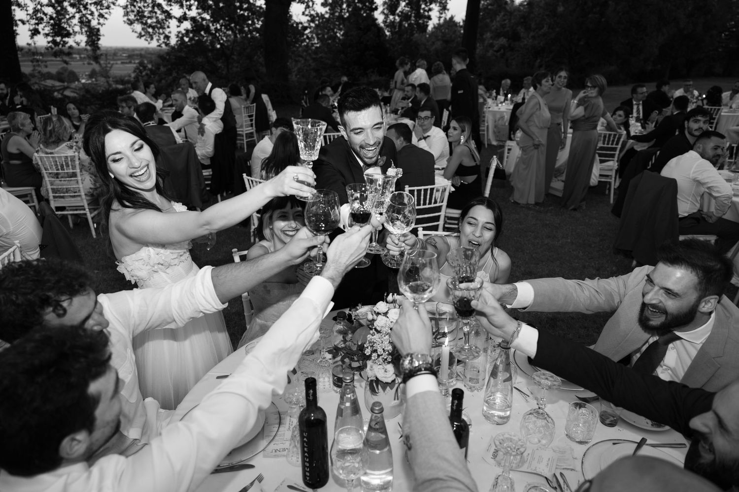 Outdoor wedding reception with guests dining at tables under trees at dusk.