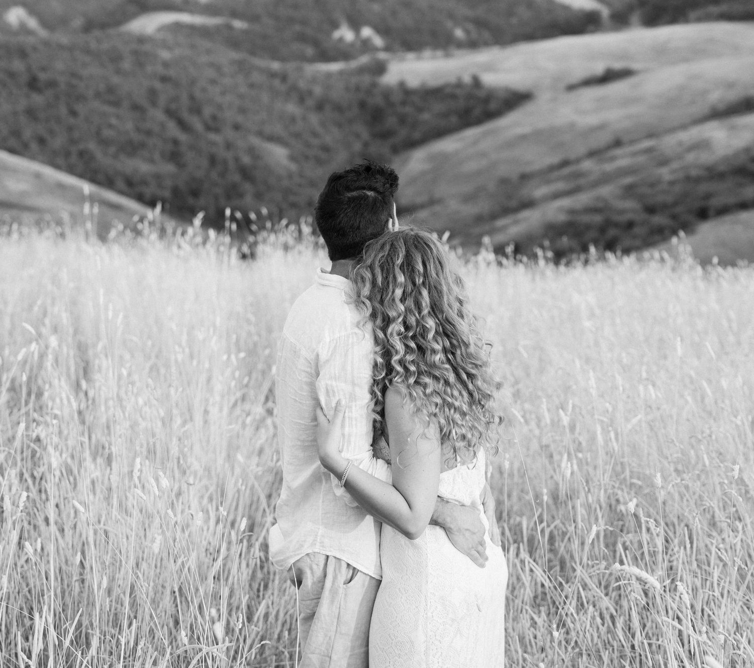 Romantic couple walking through a sunlit field of tall grass in black and white.