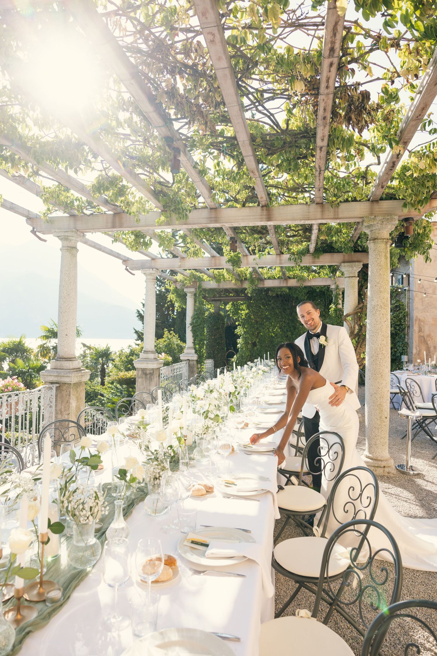 Elegant outdoor dining setup with white tablecloths and floral arrangements under a vine-covered pergola in natural sunlight.