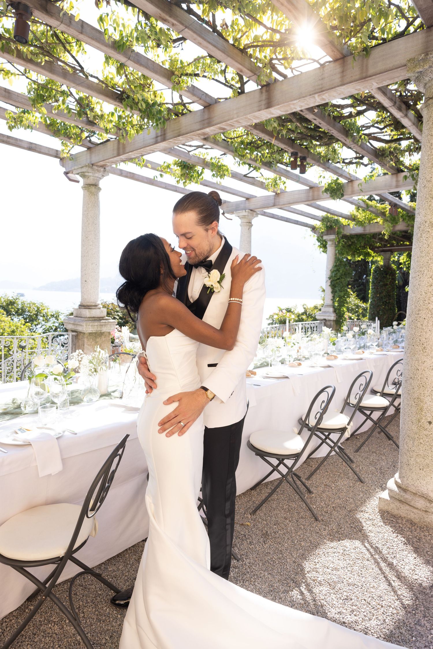 Romantic couple shares intimate dance moments under a sunlit garden pergola with white folding chairs in background.