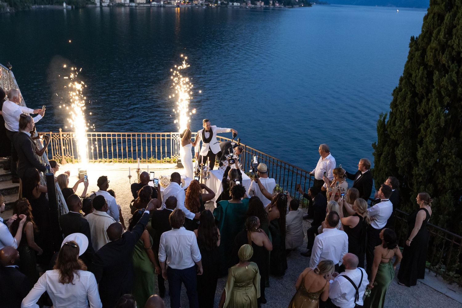 Evening celebration by a lake with sparklers illuminating a waterfront terrace as guests gather for a festive event.