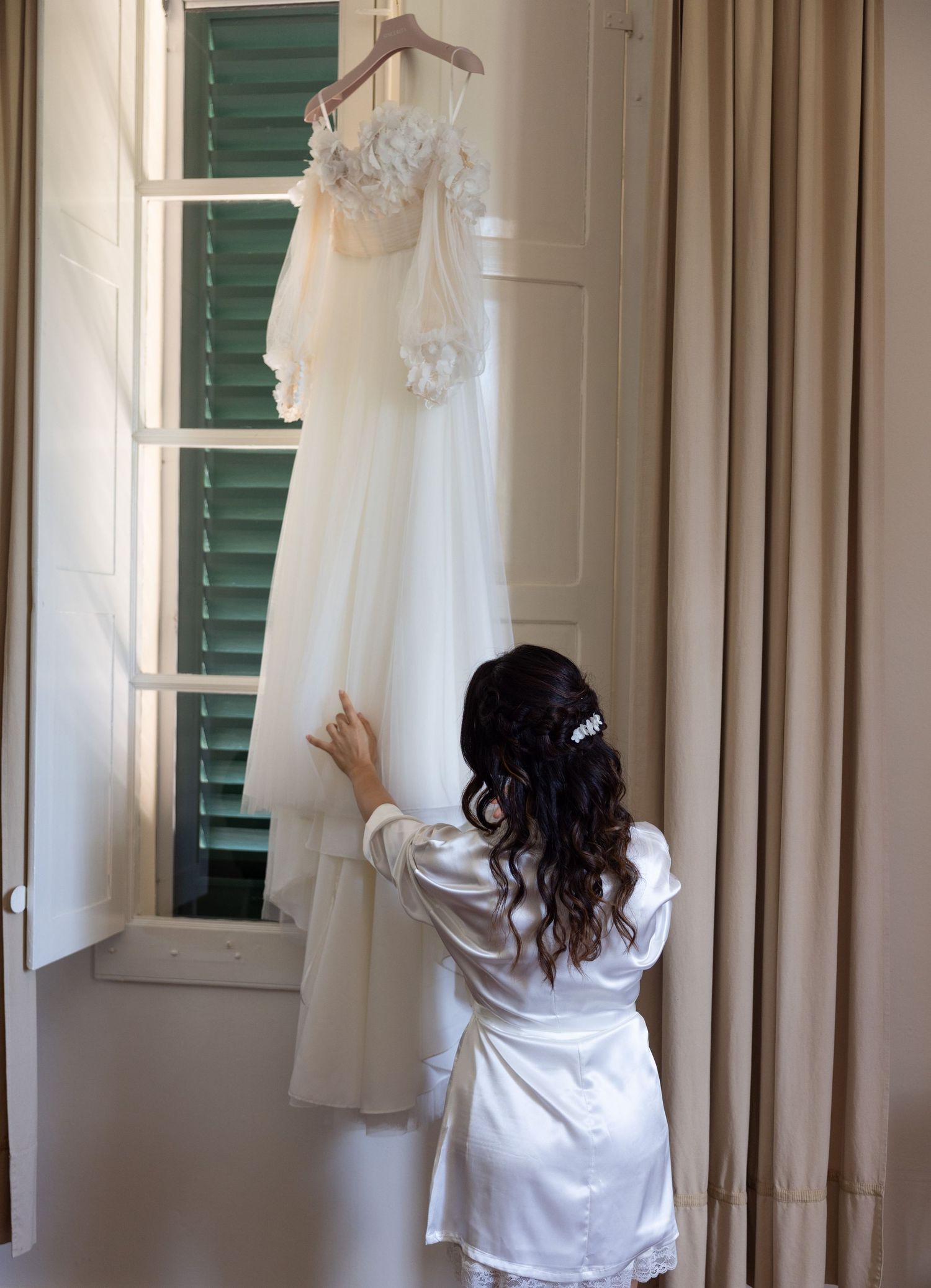 Wedding dress hanging in window frame with green shutters in natural light.