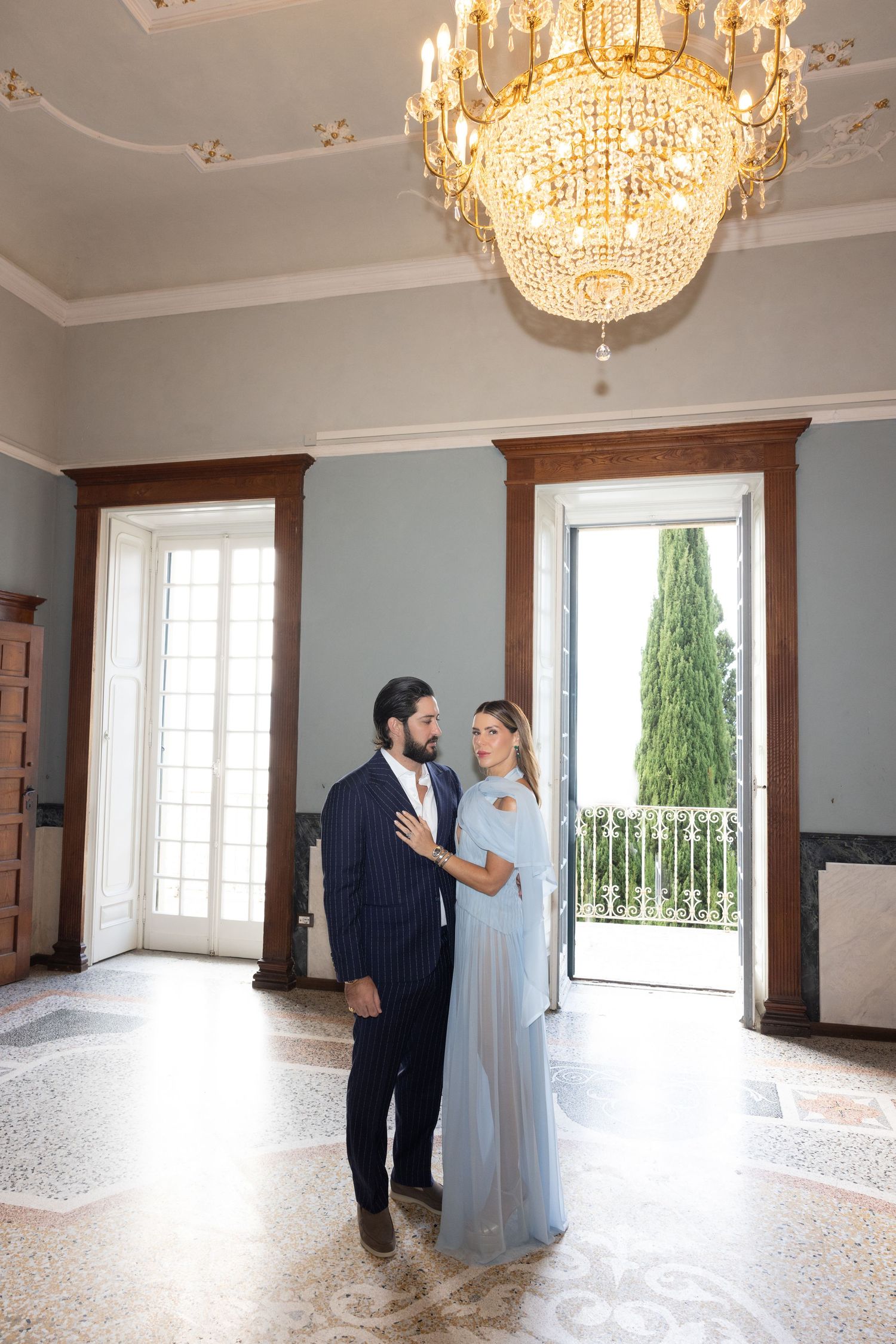A couple shares a tender moment under elegant crystal chandeliers in a bright ballroom with large windows.