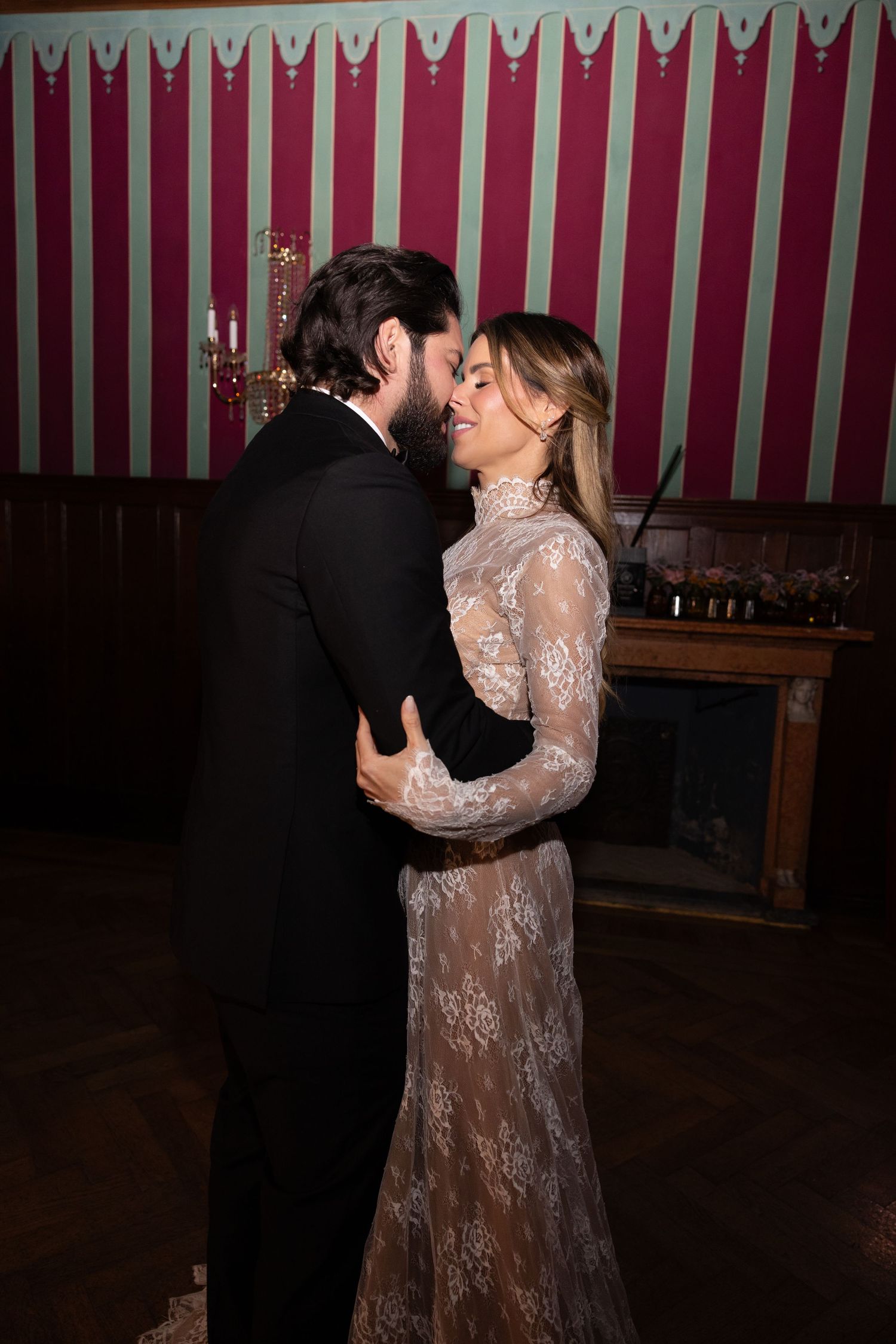 A couple in formal attire share an intimate moment against a striped burgundy wall at an elegant event.