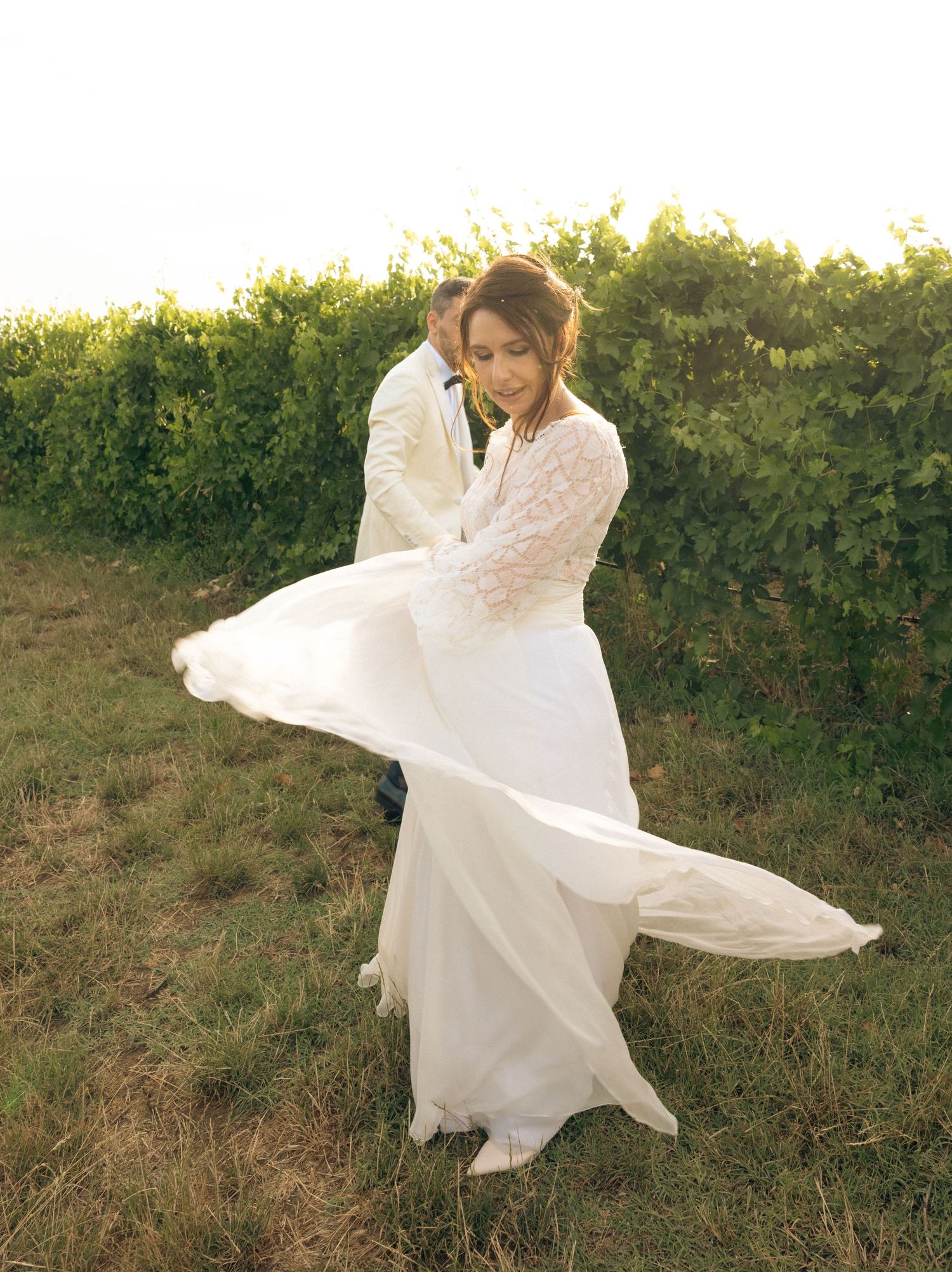 Bride in lace dress twirls playfully in a vineyard at sunset.