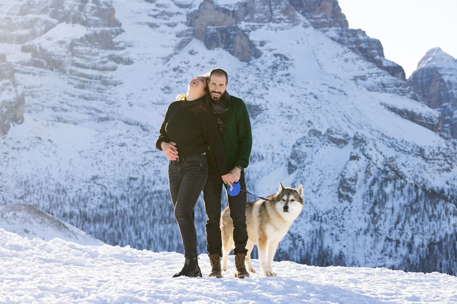 Couple standing together against snowy mountain landscape with their dog.