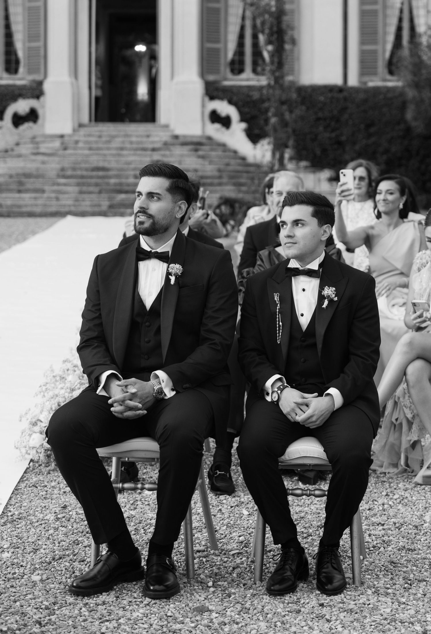 Wedding party in black tuxedos sitting casually on stone steps during an outdoor celebration.
