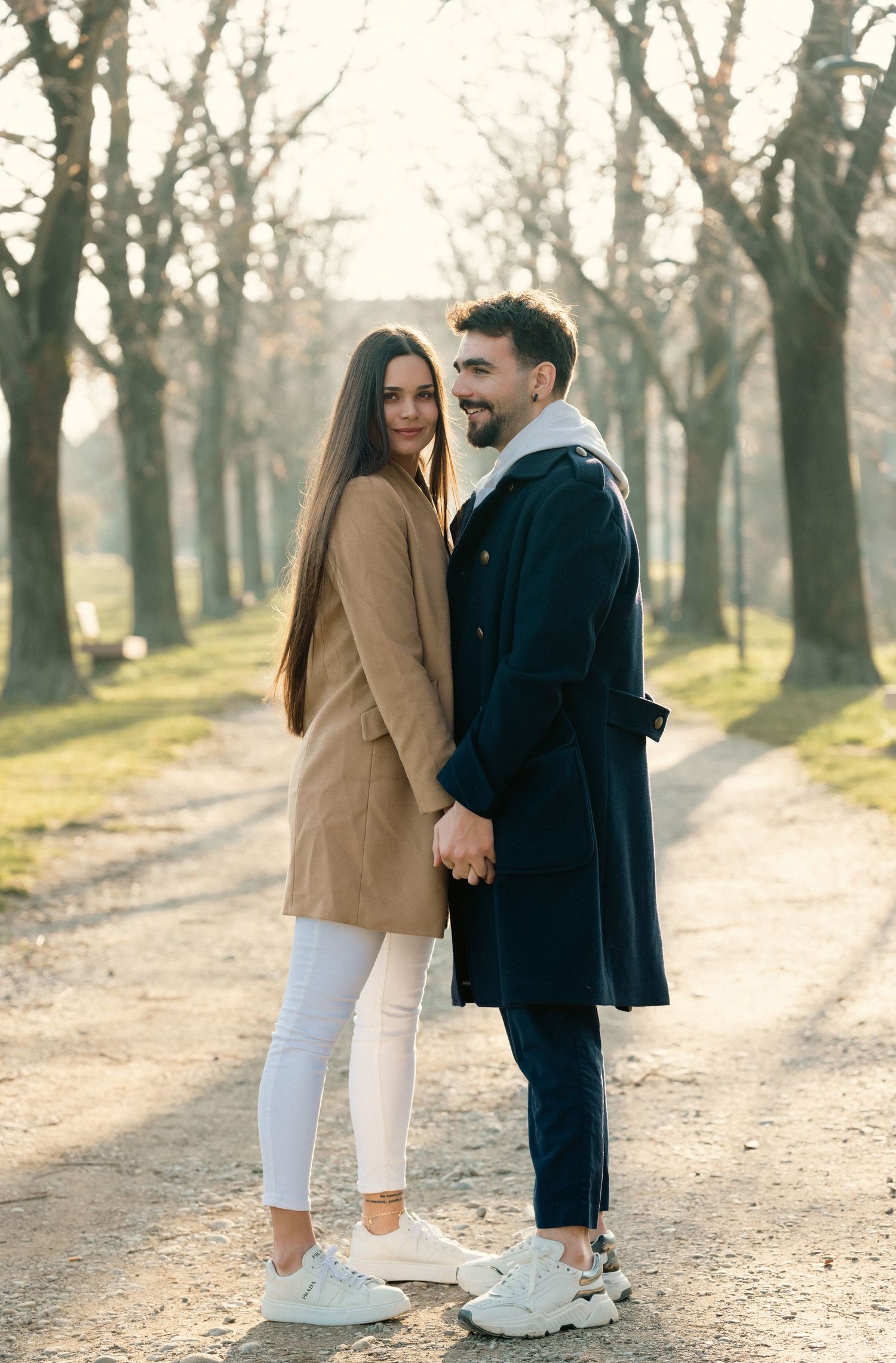 Couple walking on a tree-lined path wearing contrasting tan and black coats.