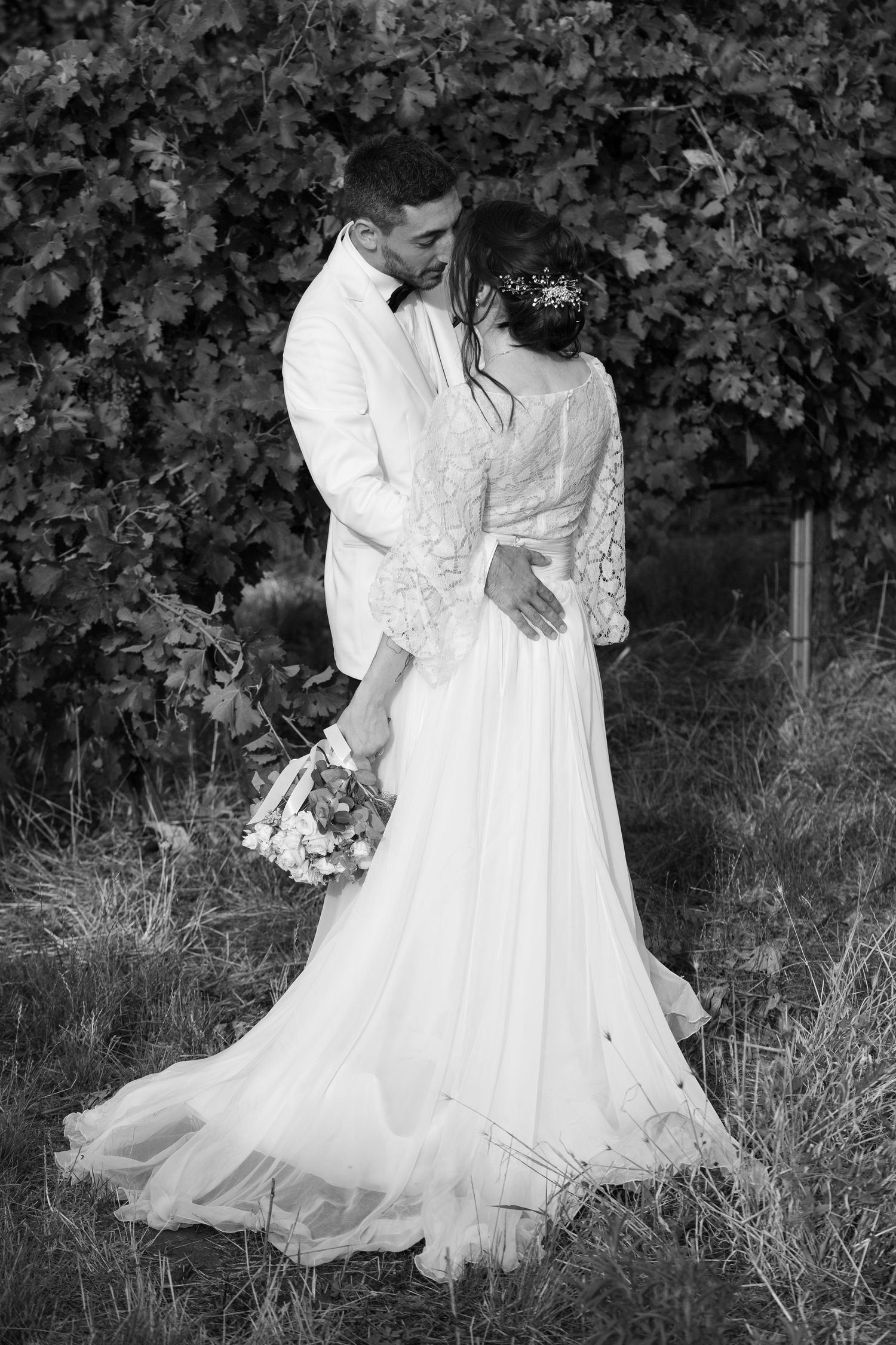Black and white portrait of newlyweds standing together against a leafy backdrop.