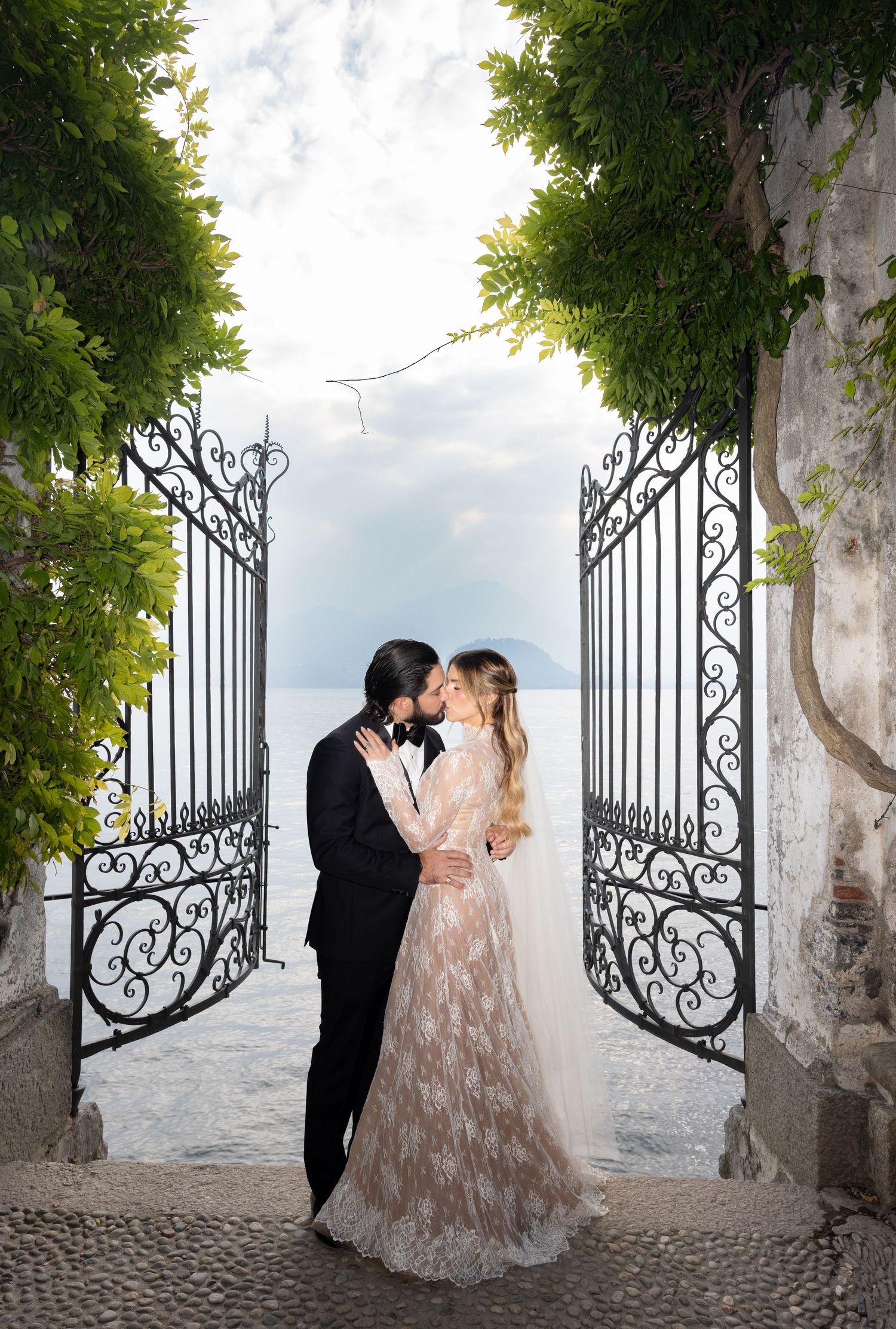 Wedding couple embracing by decorative iron gates with trees and cloudy skies in background.