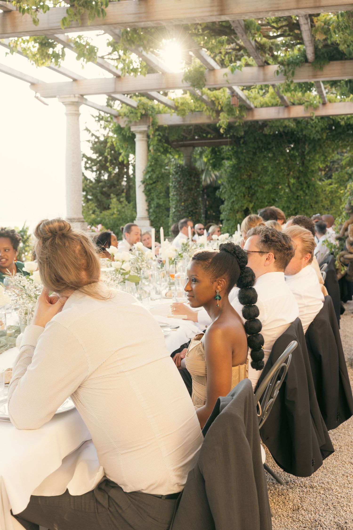 Wedding guests seated at outdoor reception tables under a white pergola with greenery overhead during sunset.