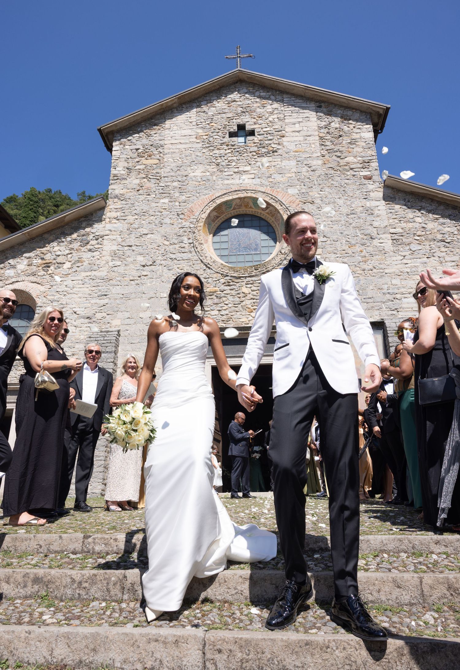 Wedding guests gather outside a rustic stone church on a sunny day during a celebration.