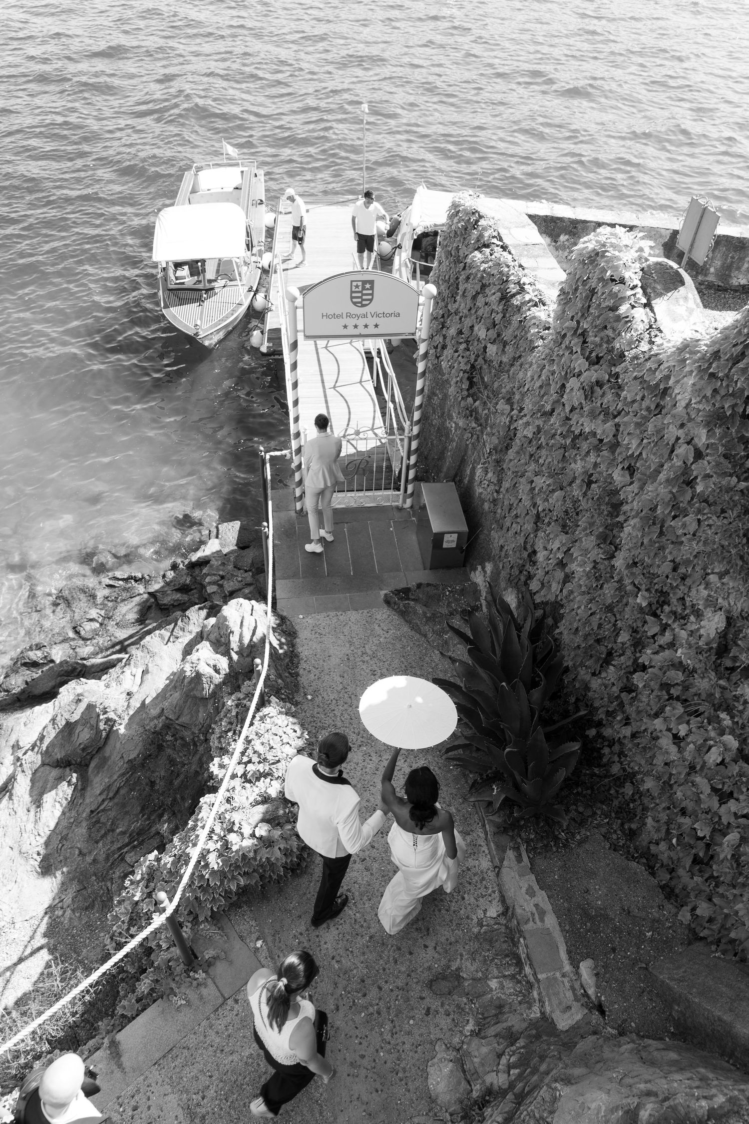 A black and white aerial view of people with parasols descending stone steps near a cliff edge by the ocean.