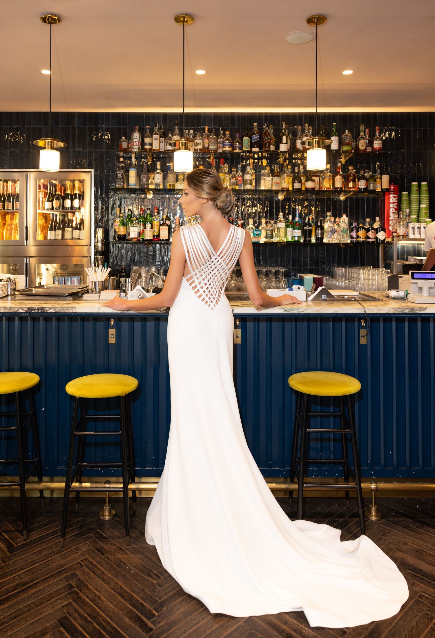 A white wedding dress displayed at an elegant bar with yellow bar stools and illuminated bottle shelves.