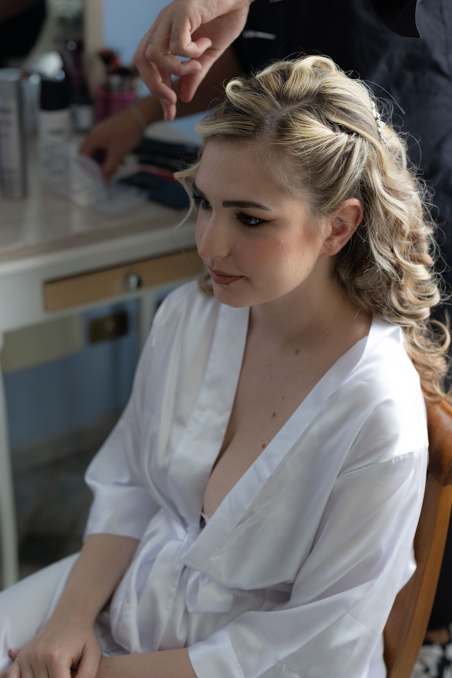 Bride getting ready in white silk robe while seated in chair during morning preparations.