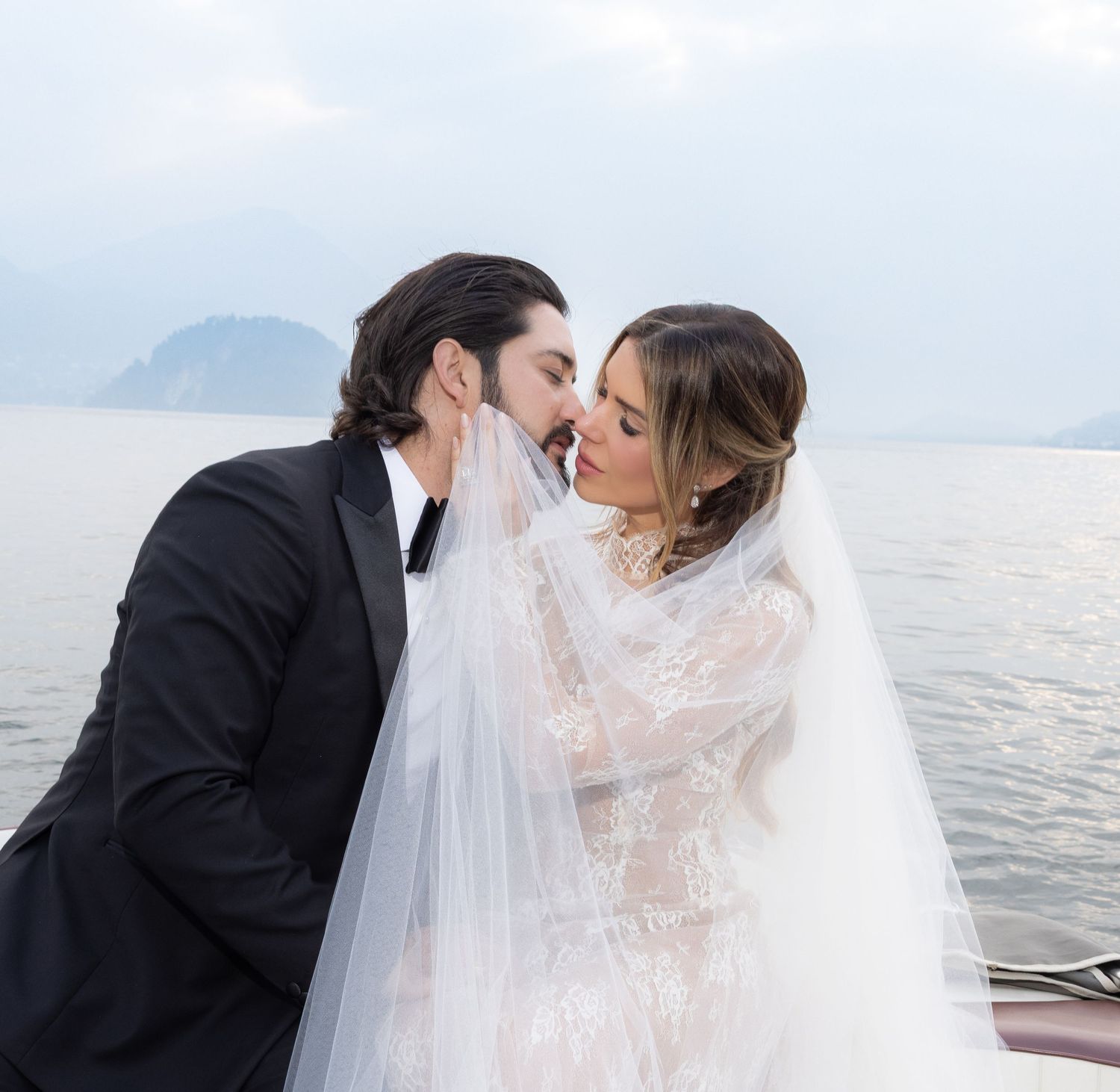 Romantic couple in wedding attire sharing intimate kisses by a scenic waterfront with mountains in the background.