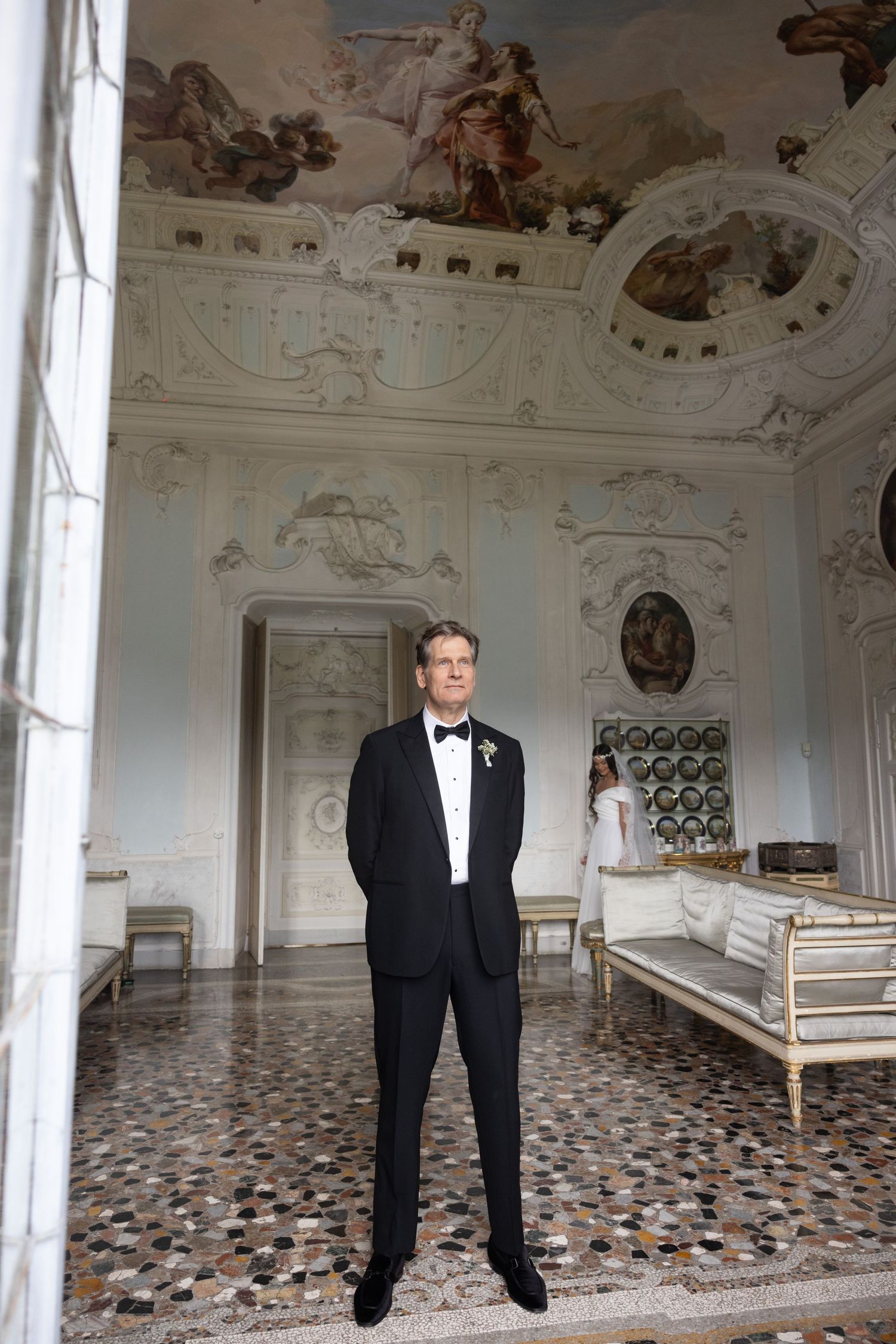 A groom in a black tuxedo stands in an ornate room with frescoed ceilings and patterned floors.