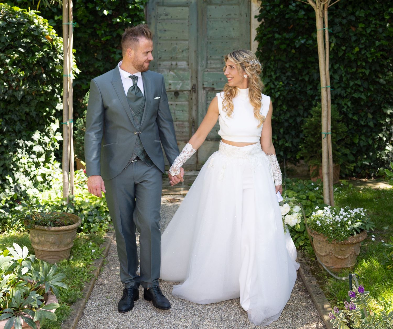 A couple in wedding attire walks along a garden path surrounded by greenery.