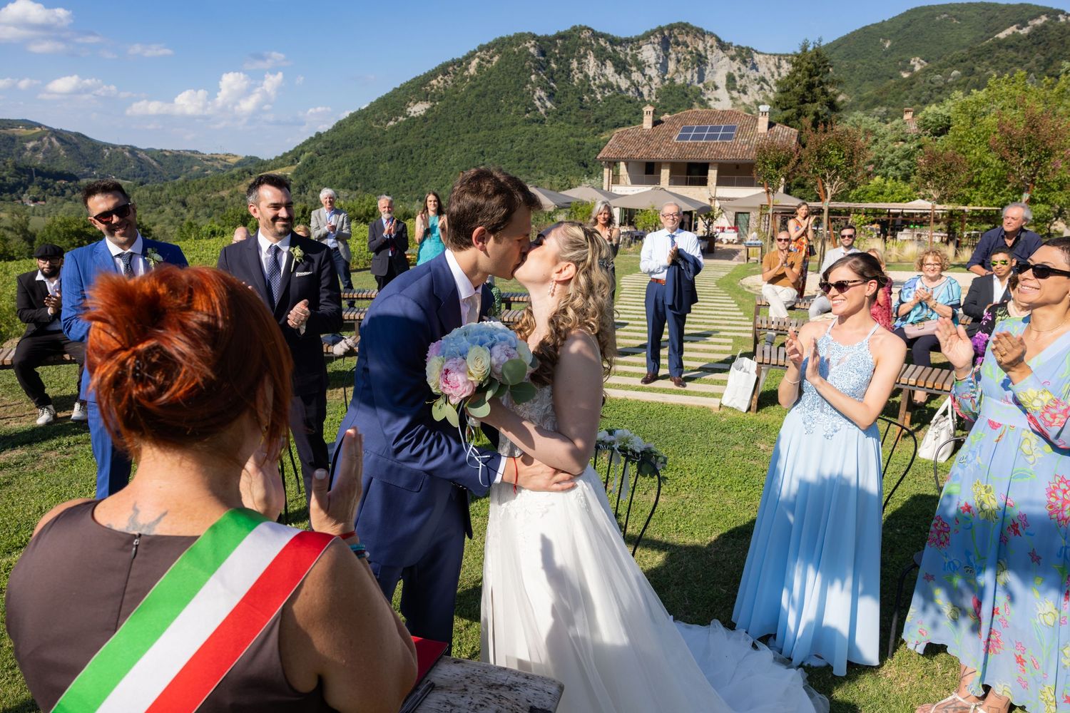 An outdoor wedding ceremony takes place in a scenic mountain setting with guests watching.