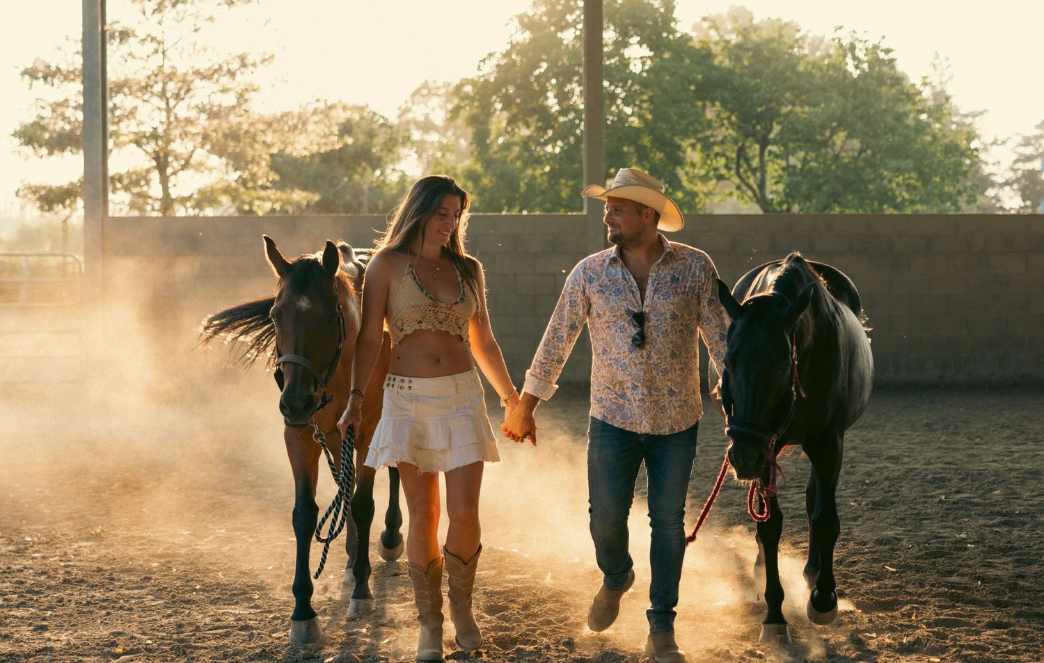 Silhouettes of people and horses walking together at sunset in a dusty arena.
