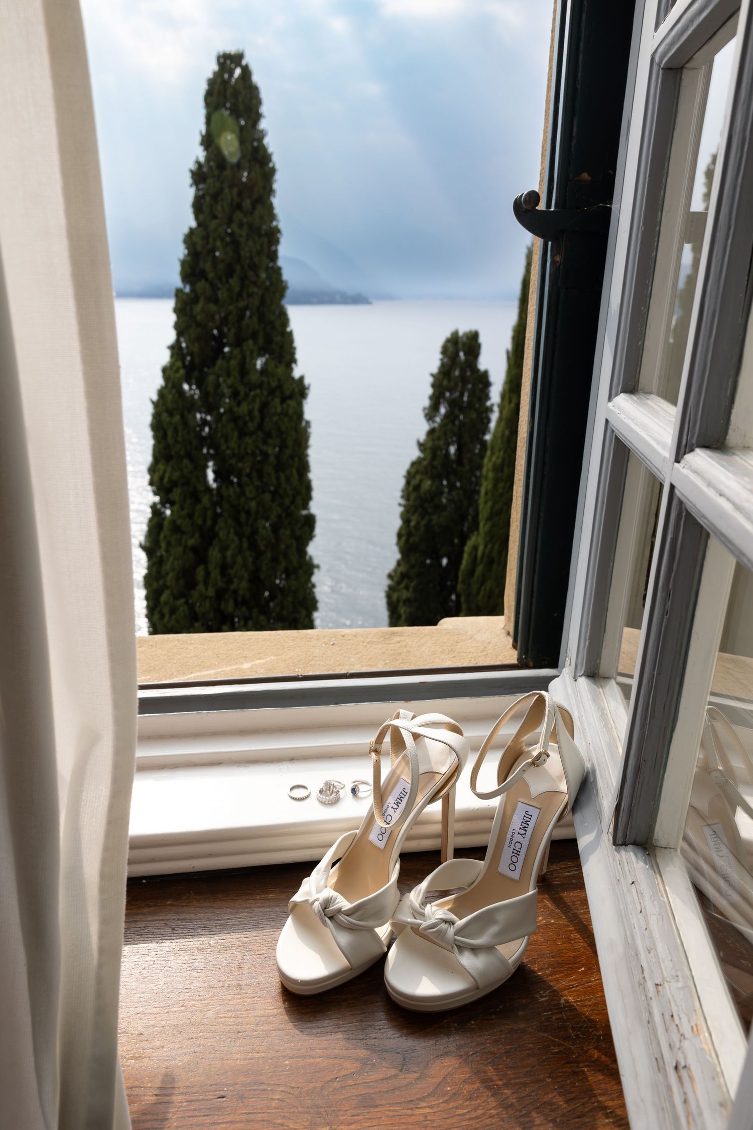 White wedding shoes Jimmy Choo sit on a windowsill overlooking a scenic lake view with cypress trees framing the scene.