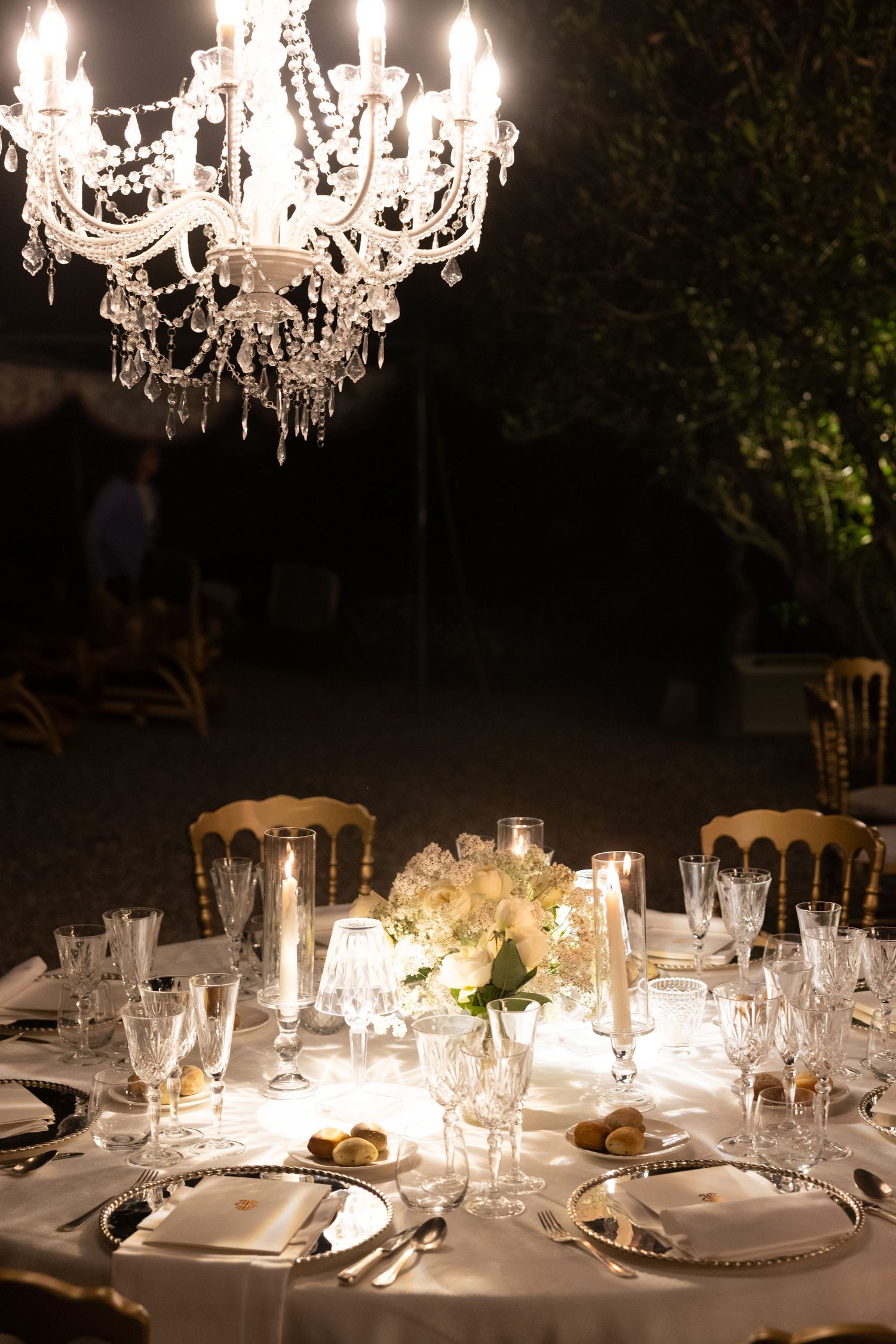 Crystal chandelier hanging over formal table settings with wine glasses and white linens.
