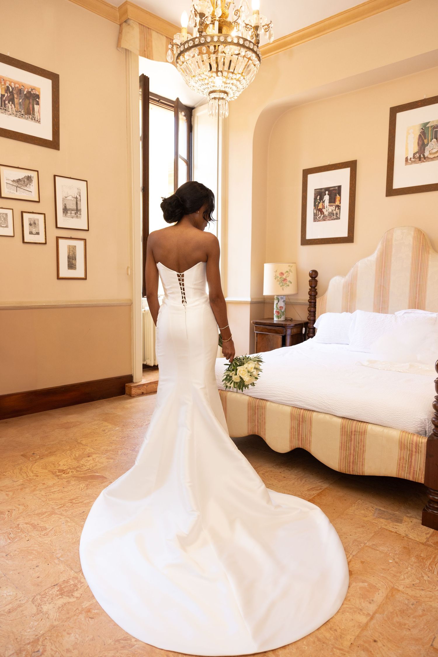 A bride in a flowing white strapless wedding gown twirls in an elegant room with a crystal chandelier and framed artwork.