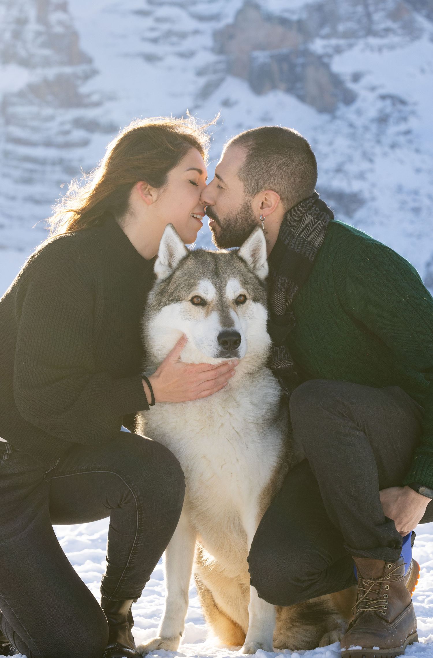 Husky dog sits between two people in the snow with mountains in background.