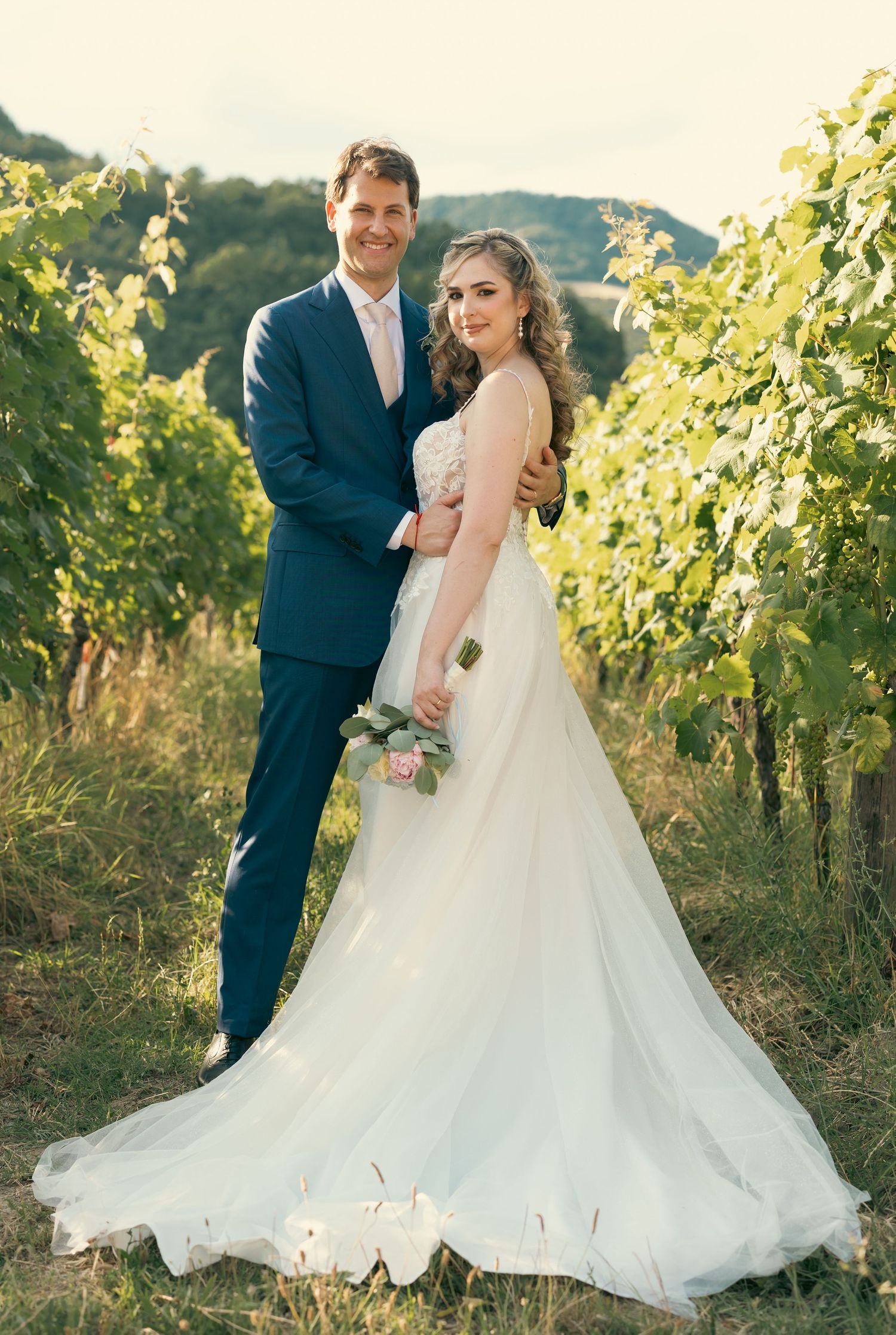 Romantic wedding couple poses in vineyard with flowing white dress and navy suit at sunset.