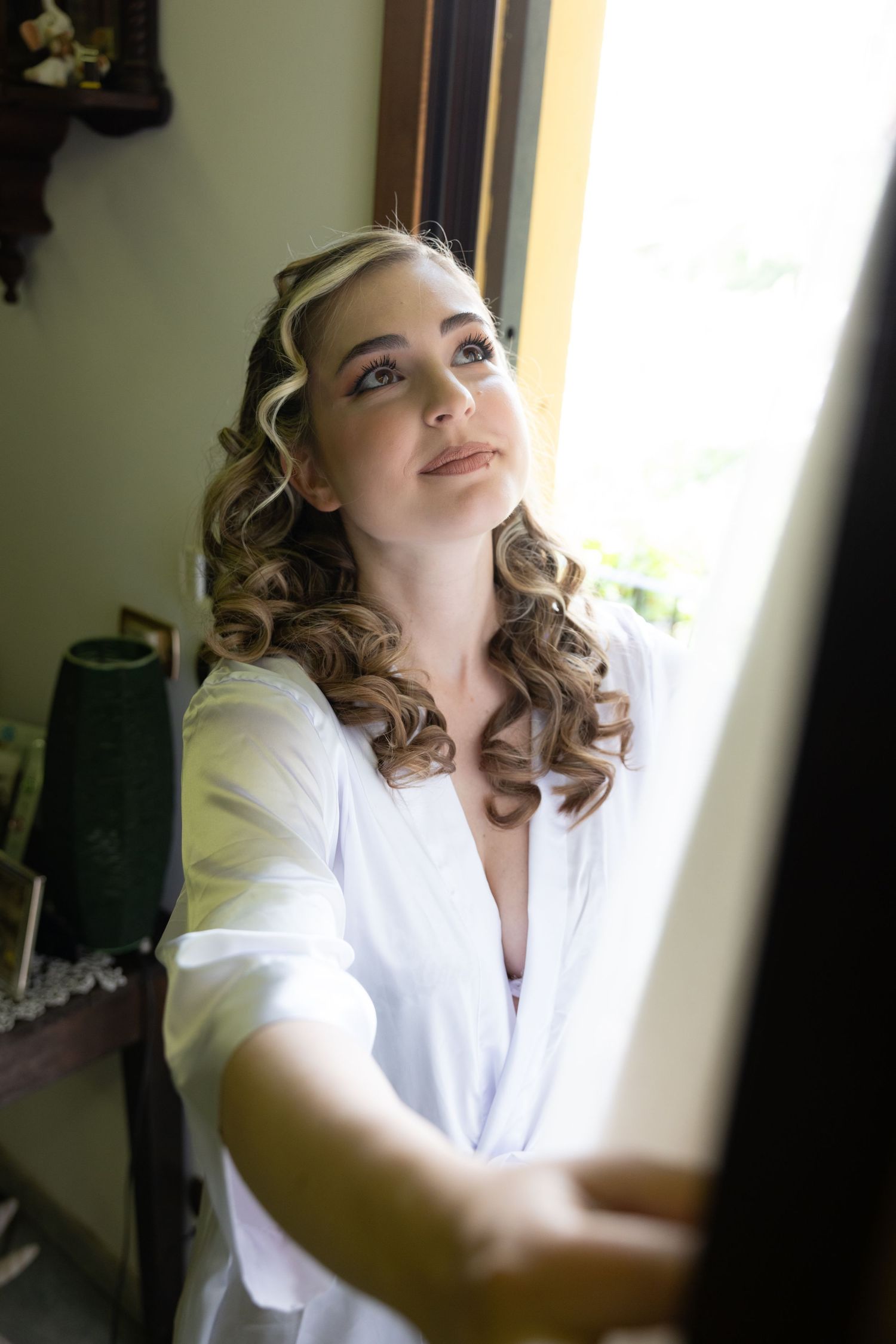 A person in a white robe getting ready by a window during wedding preparations.